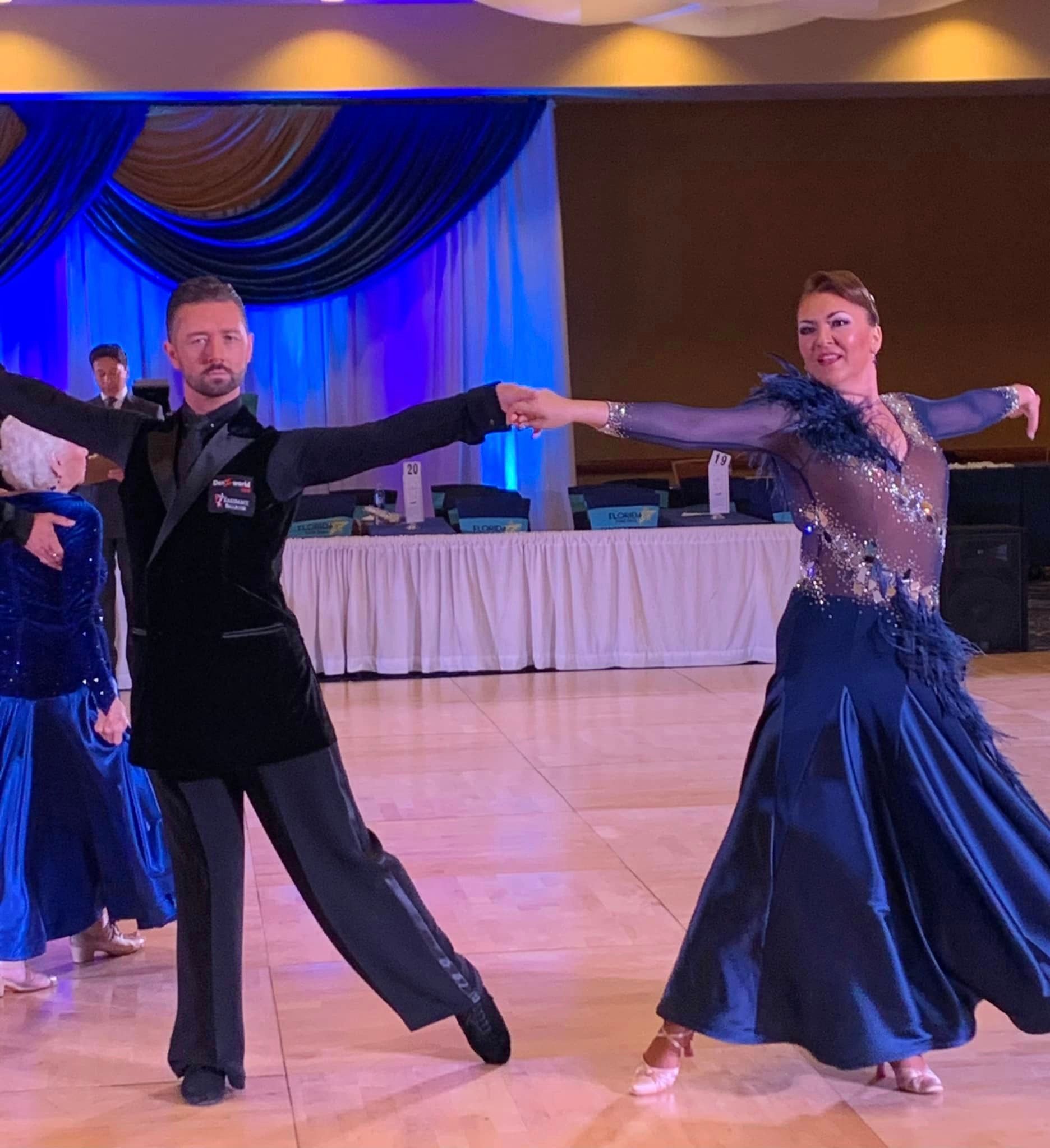 A couple dancing in a ballroom. The woman is in a blue gown, and the man is in a tuxedo.