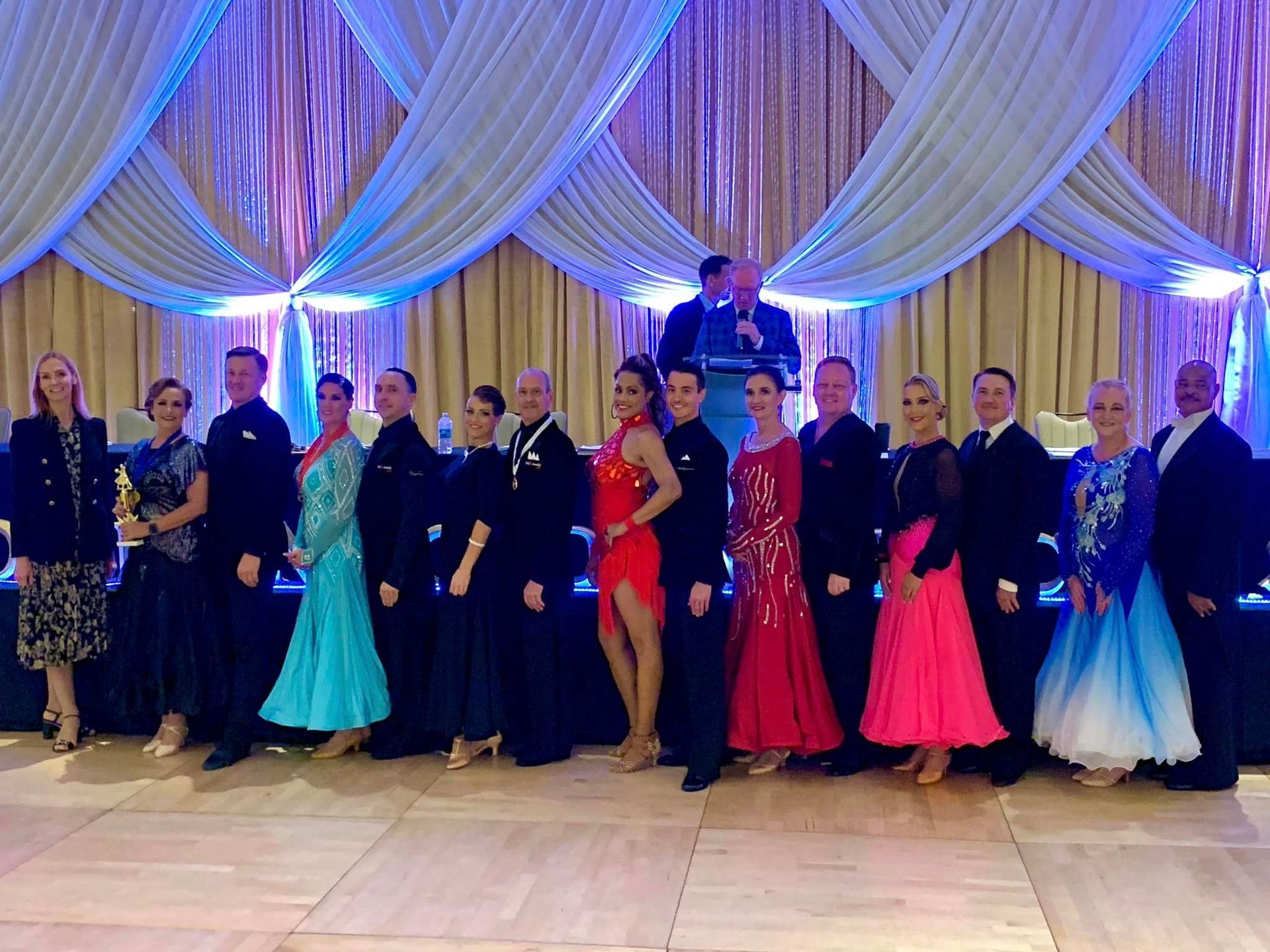 Group of ballroom dancers posing, with a man on stage, in a decorated hall.