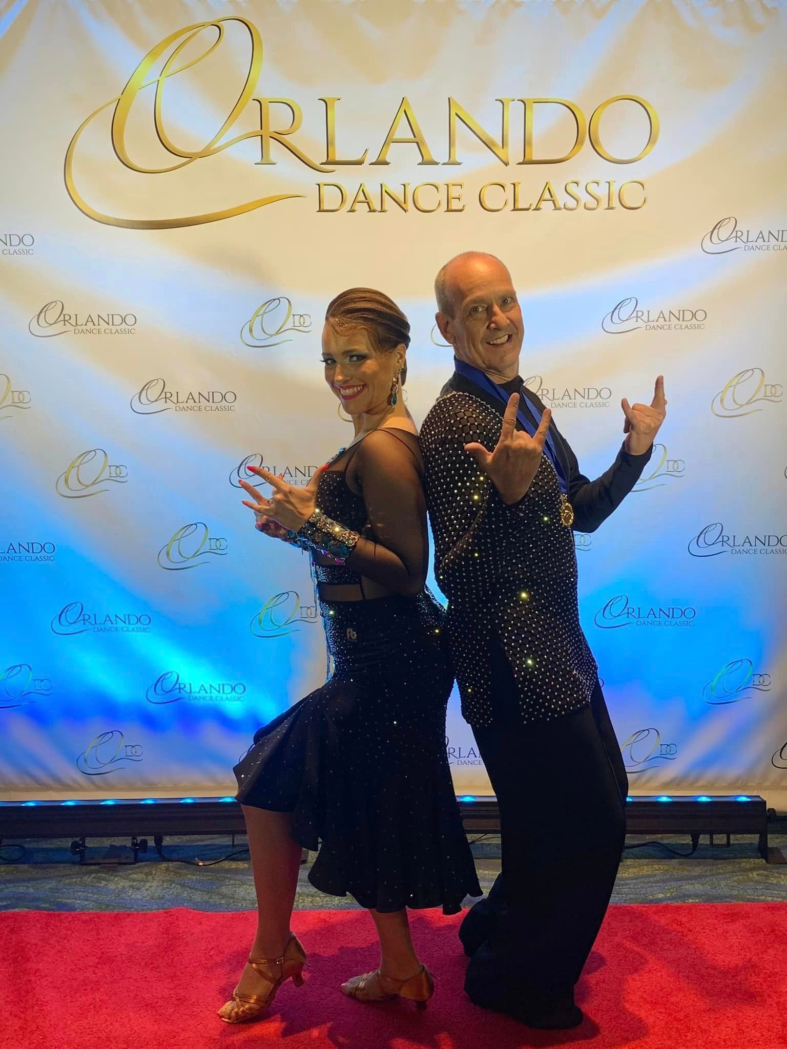Dancers pose on a red carpet at Orlando Dance Classic. Woman in black dress, man in sparkly shirt, smiling, throwing hand signs.