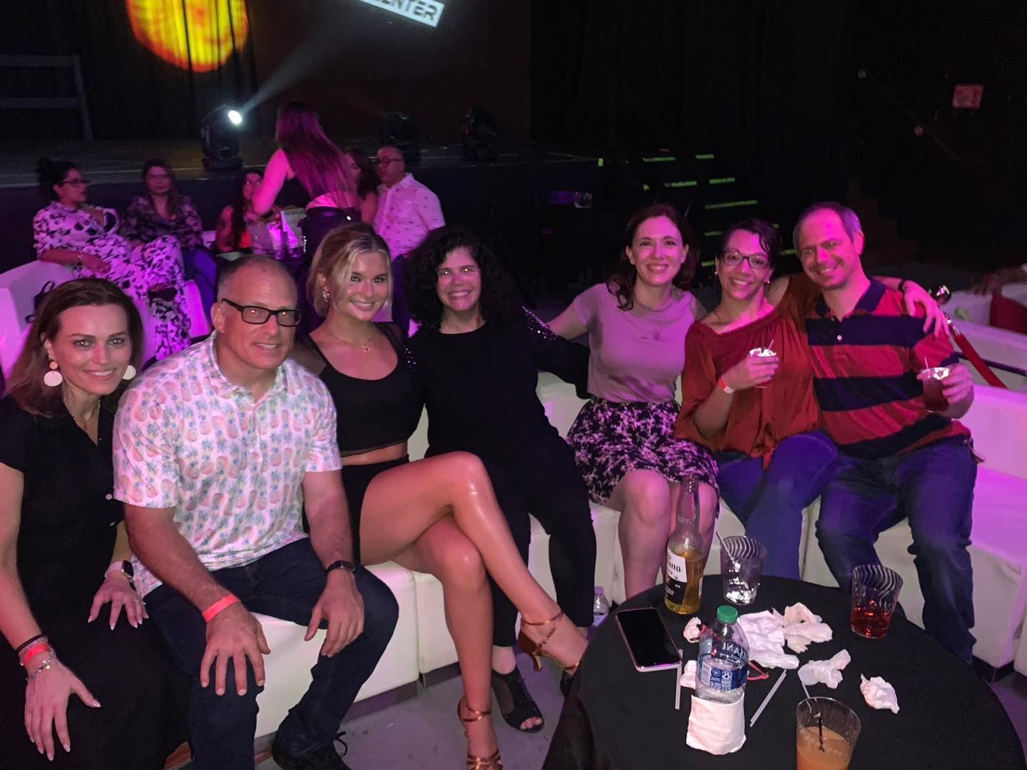 Group of people, including diverse genders and ethnicities, smiling at a party, some with drinks, sitting on a white sofa.