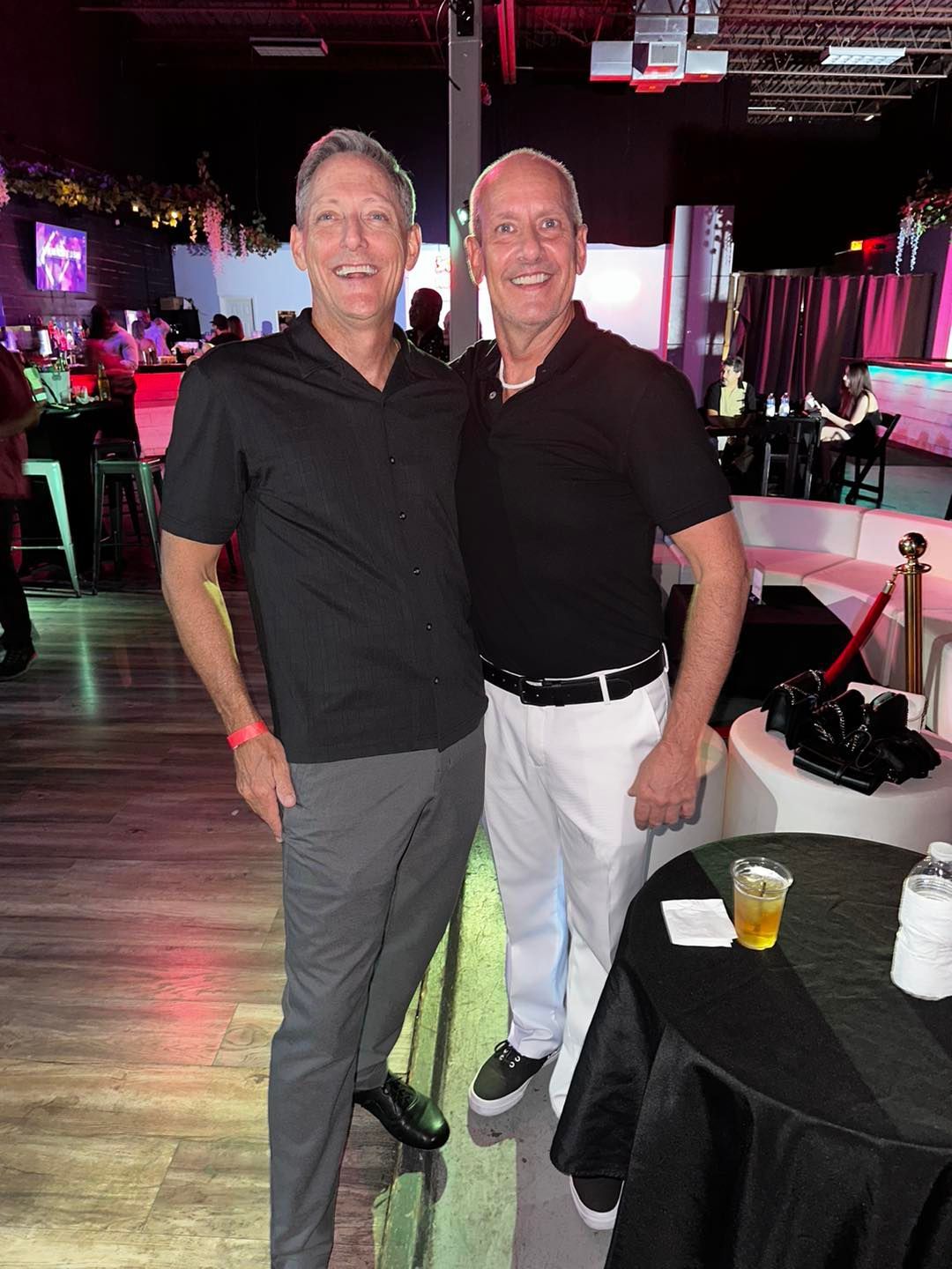 Two men smiling in a nightclub, one in black shirt and gray pants, the other in black shirt and white pants.