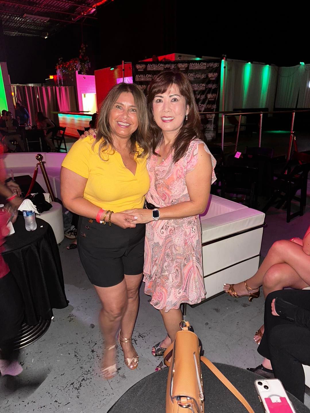 Two women smiling, embracing at a party. One in yellow shirt and shorts, the other in a pink dress.