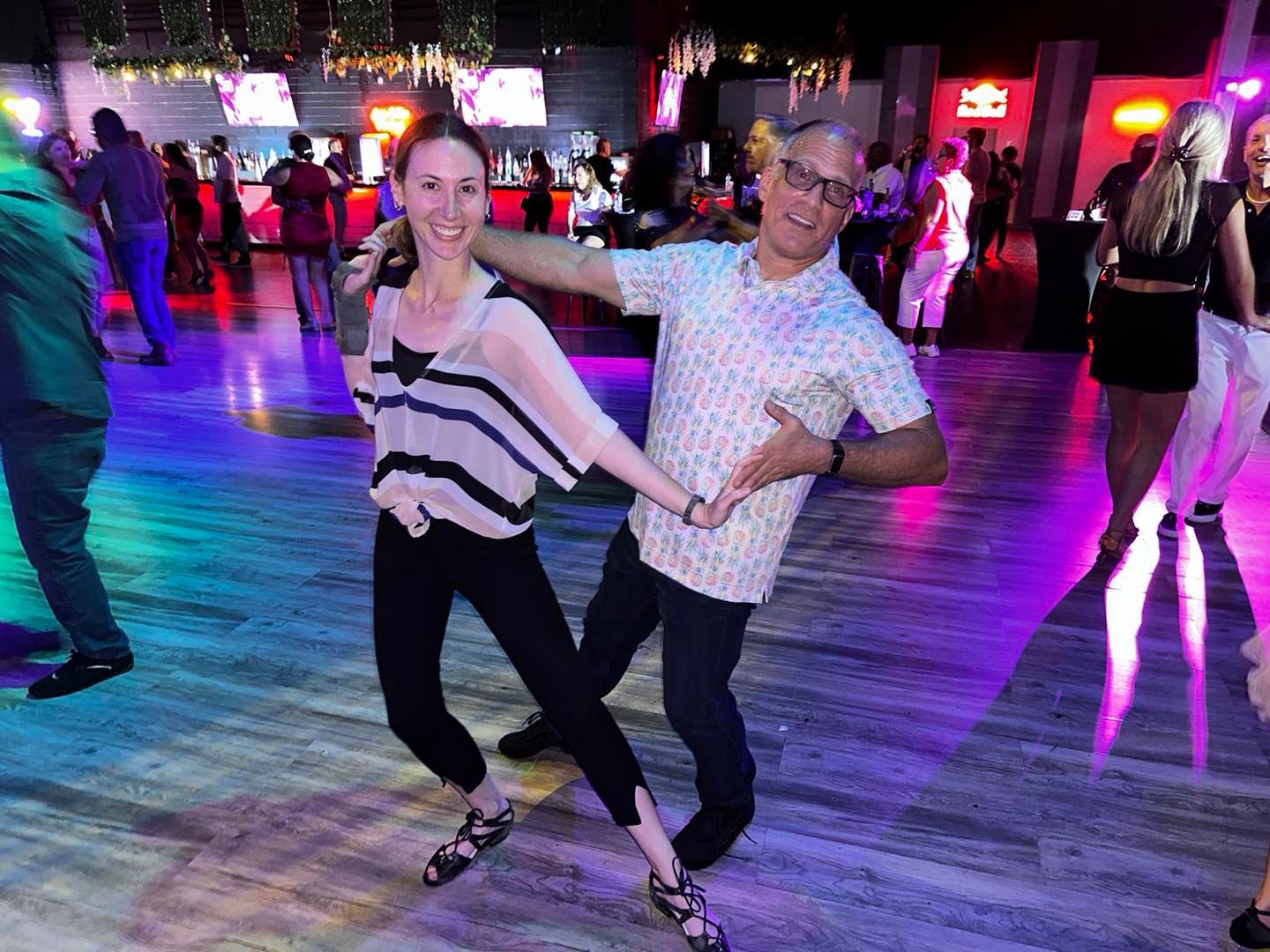 Man and woman dancing on a colorful lit dance floor. Man has a playful expression while gesturing, and woman smiles.