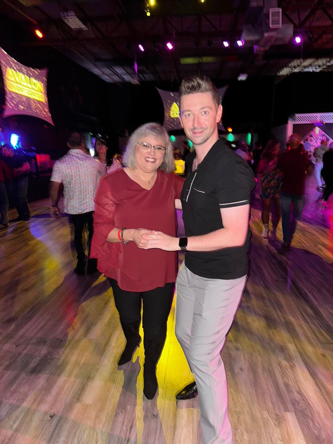 Woman and man smiling while dancing on a wooden dance floor. The woman is in a red top and black pants. The man is wearing a black shirt and grey pants.