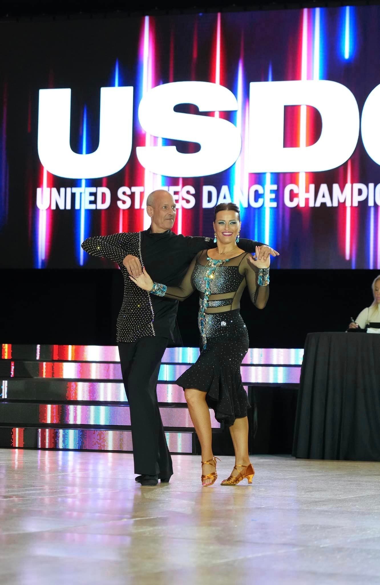 A couple ballroom dances on a stage, with the US Dance Championship logo in the background.
