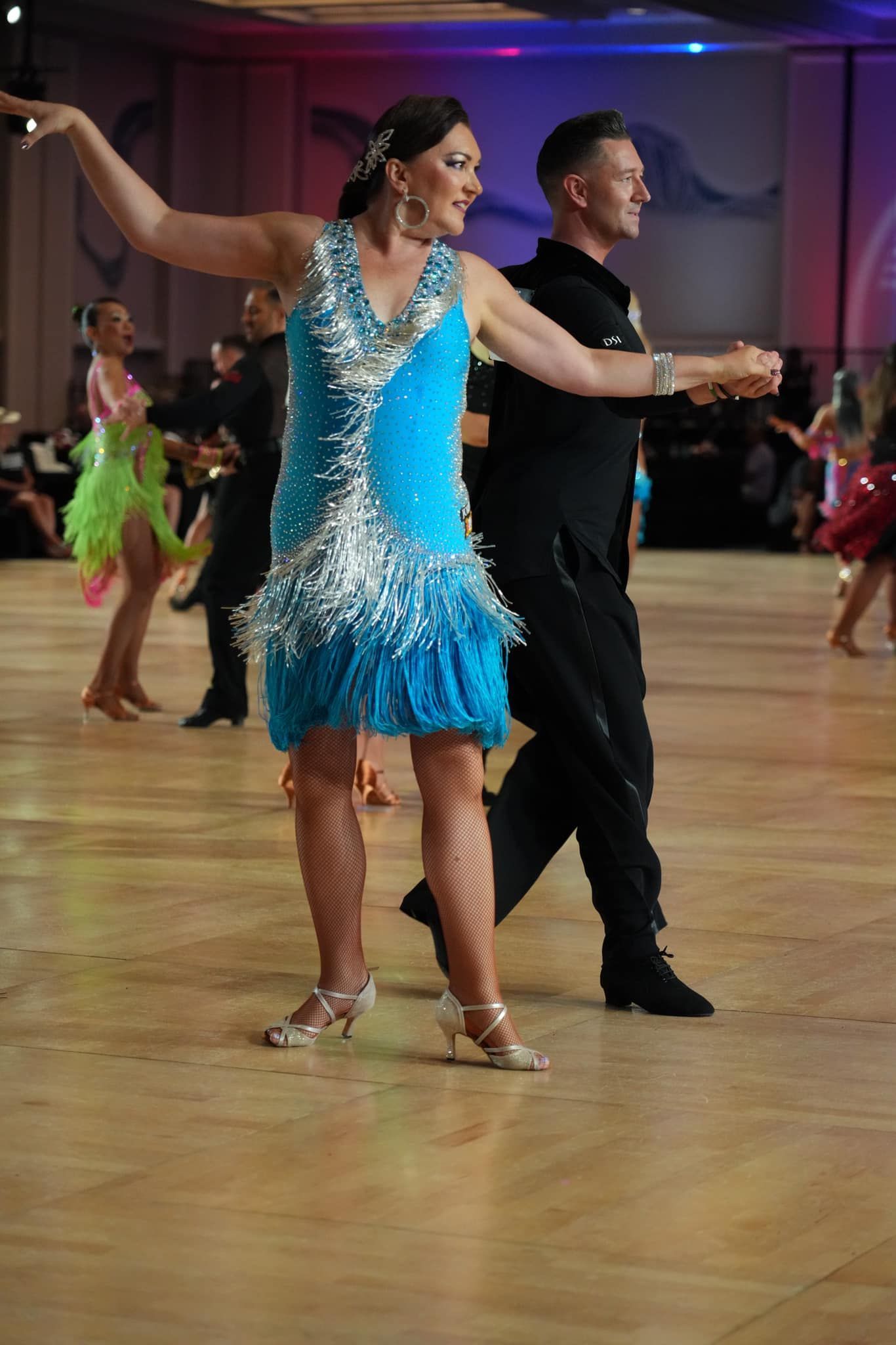 Woman in turquoise dress dances with a man in a black outfit on a ballroom dance floor.