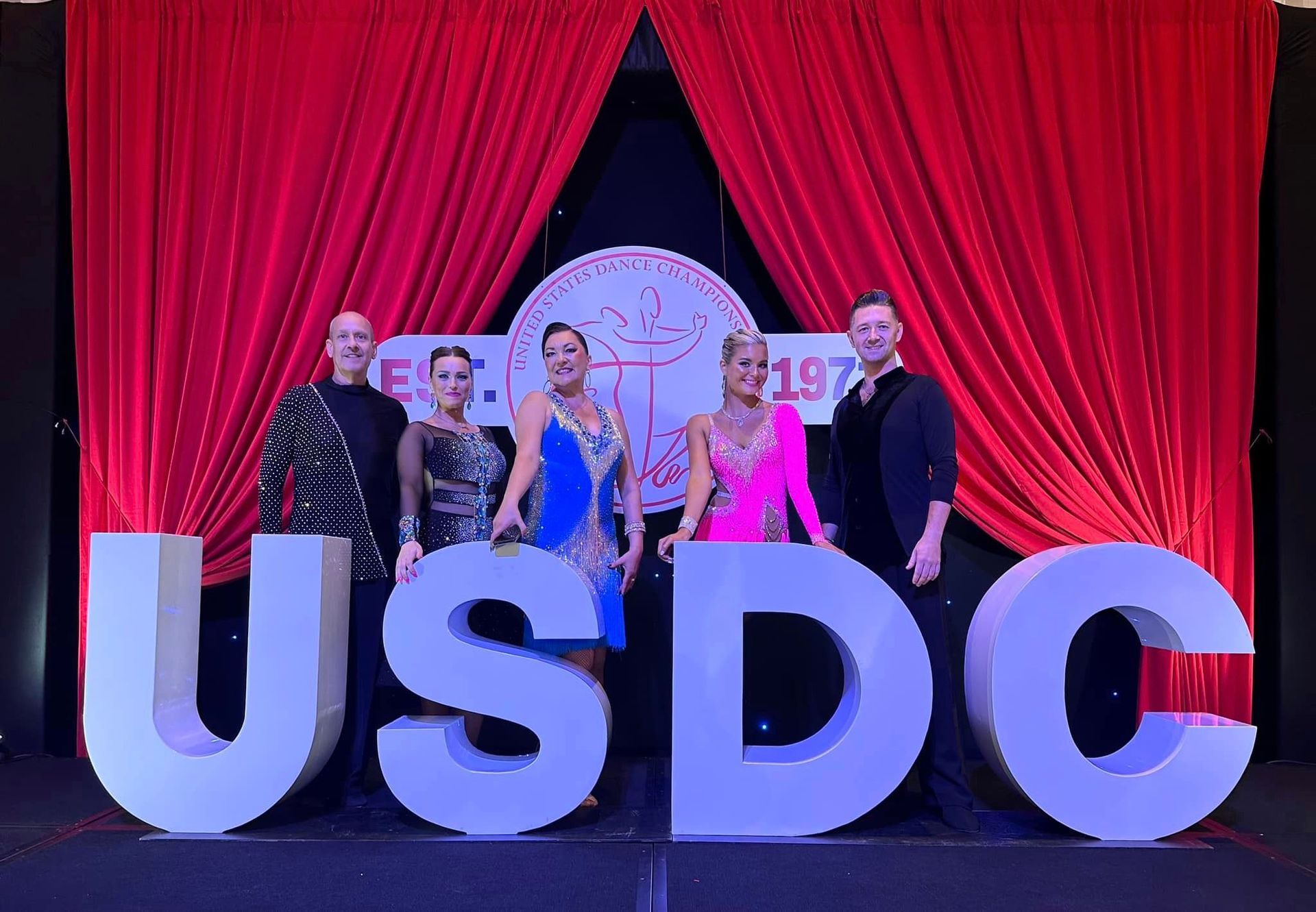 Dancers pose behind the USDJ logo at a ballroom event. Red backdrop; colorful costumes.