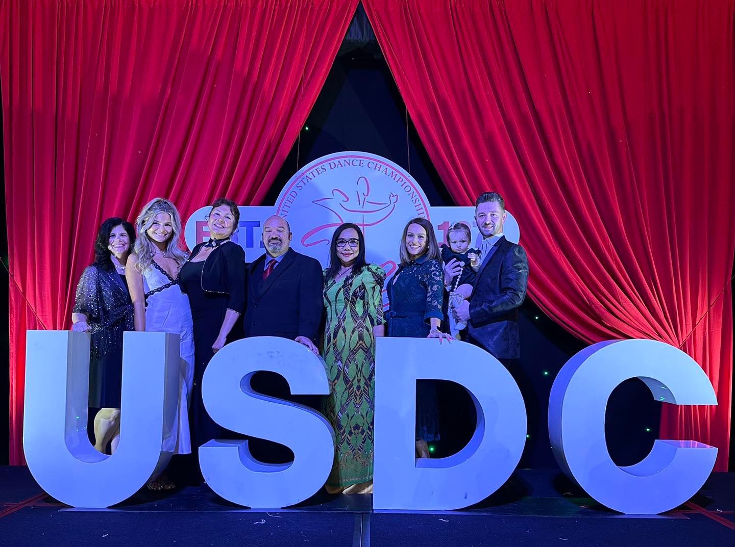 Group of people posing at a formal event with “USDC” letters in front of a red curtain backdrop.