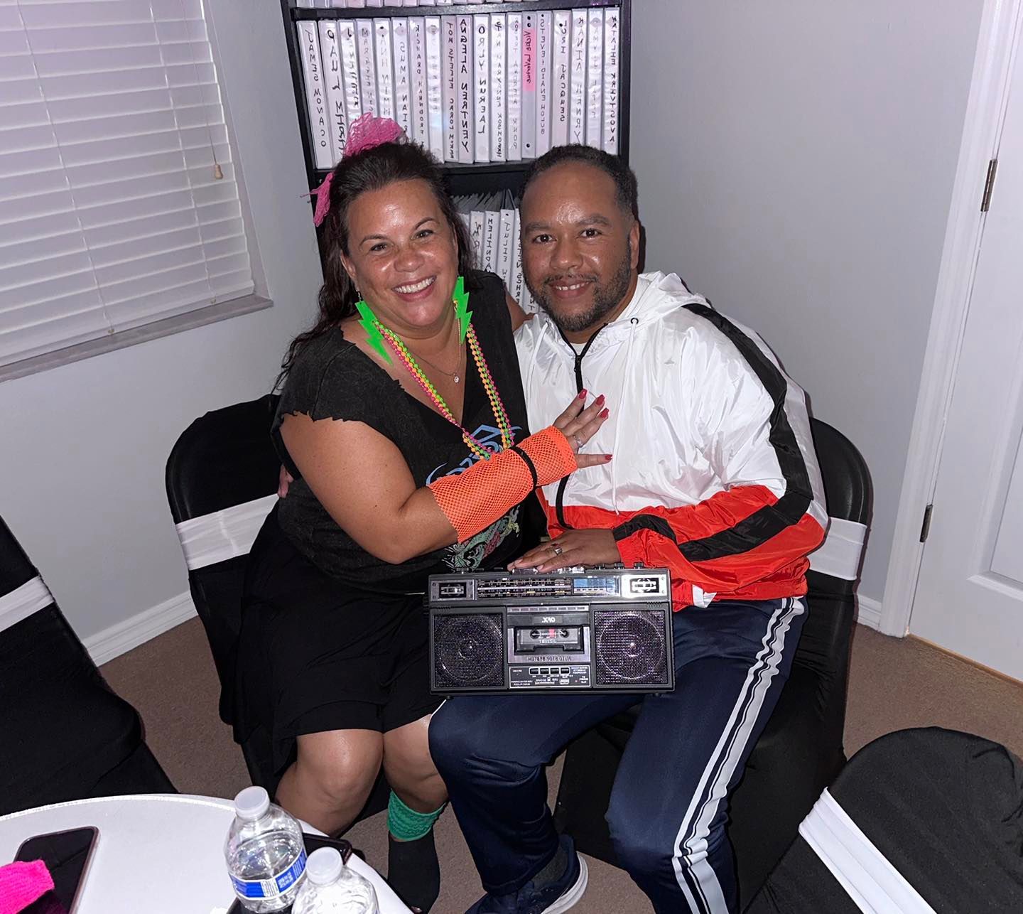 Two people in 80s attire pose with a boombox; woman has pink headband, man wears a jacket; indoors.