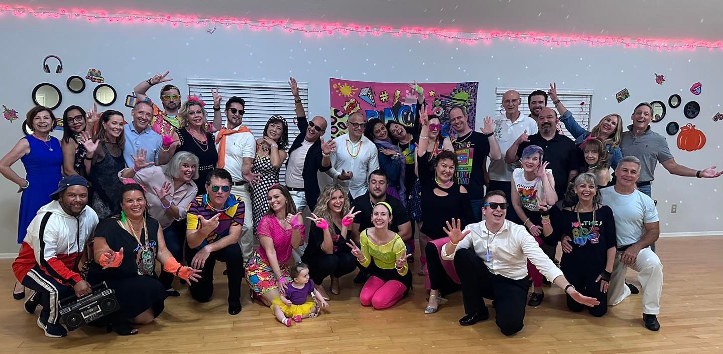 Group of people in 80s costumes pose in a dance studio, some in neon outfits.