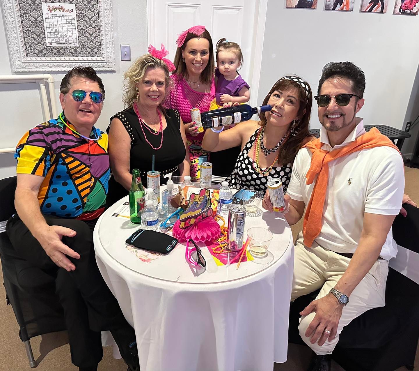 Group of people in colorful 80s attire at a table with drinks, smiling, and posing.