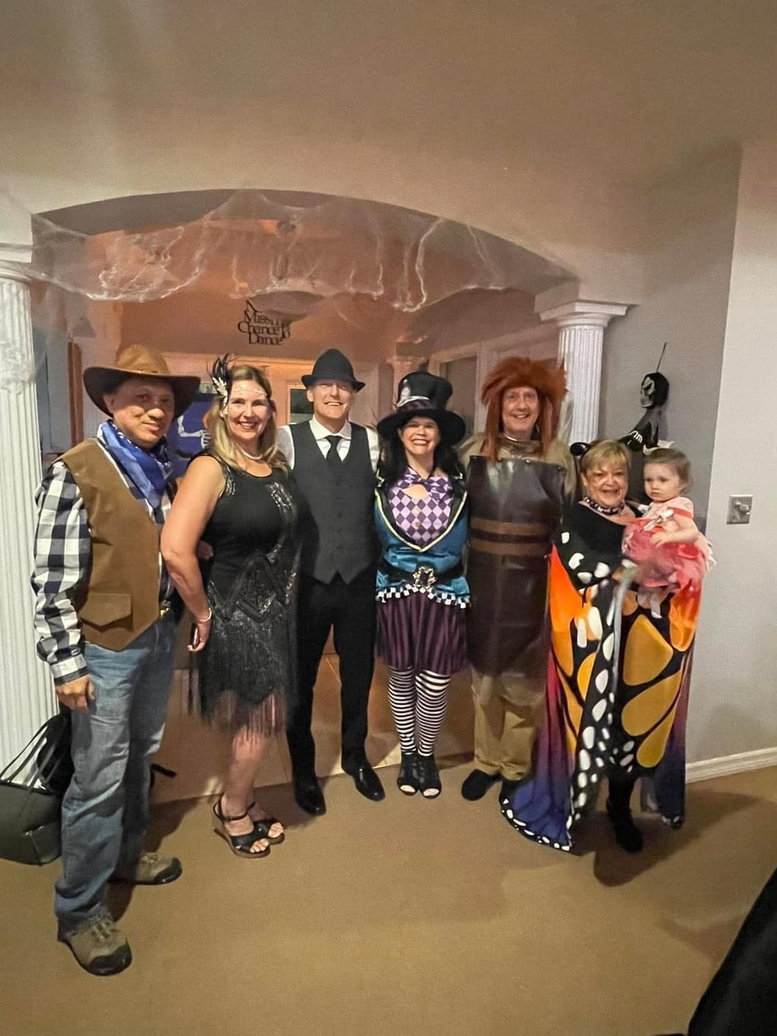Group of people in costumes pose indoors. Cowboy, flapper, gangster, witch, knight, butterfly and child.