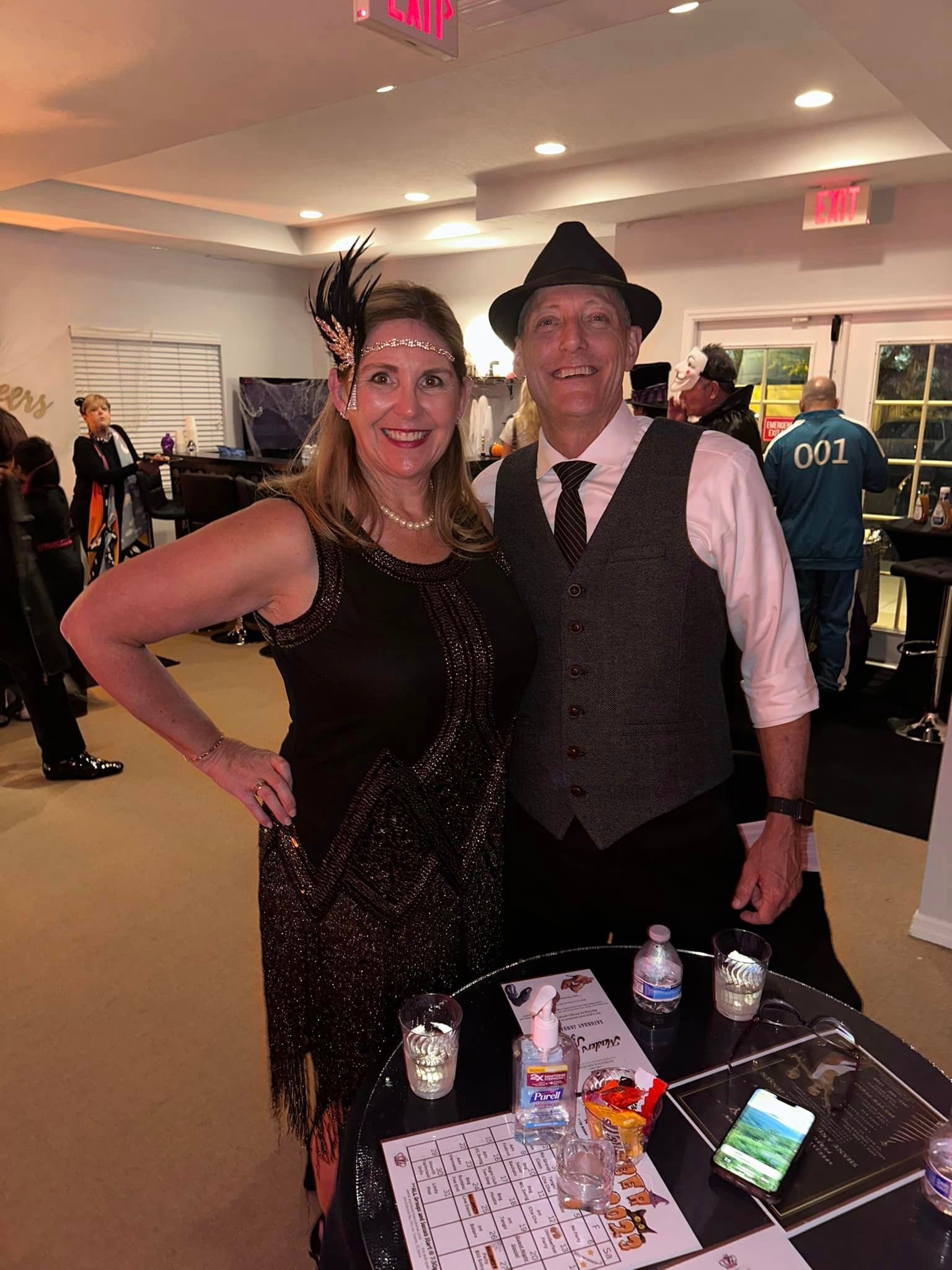 Couple in Gatsby attire at a party. Woman in sparkly dress, man in vest and hat, standing near a table.