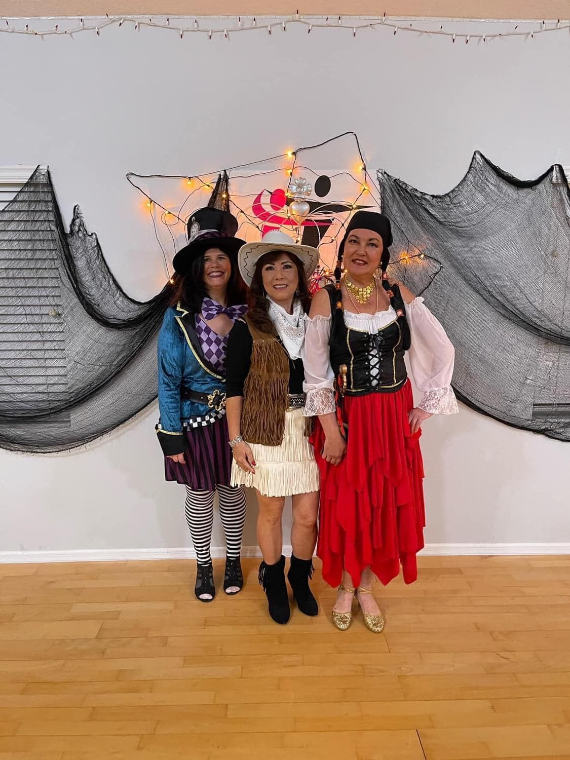 Three women in Halloween costumes pose in front of a decorated wall.