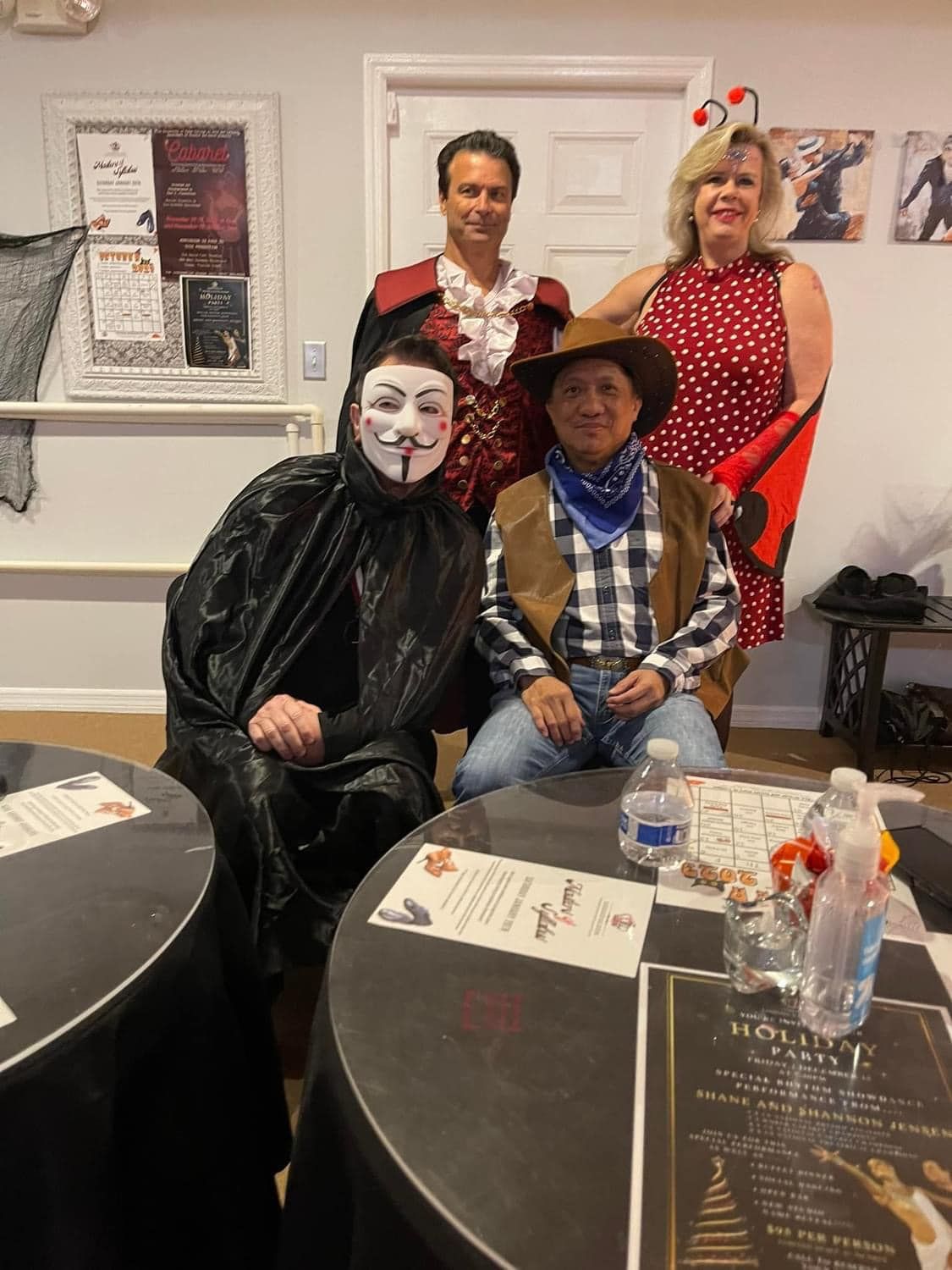 Group of four people in costumes at a gathering. One wears a mask, cowboy, vampire, and polka dot dress.