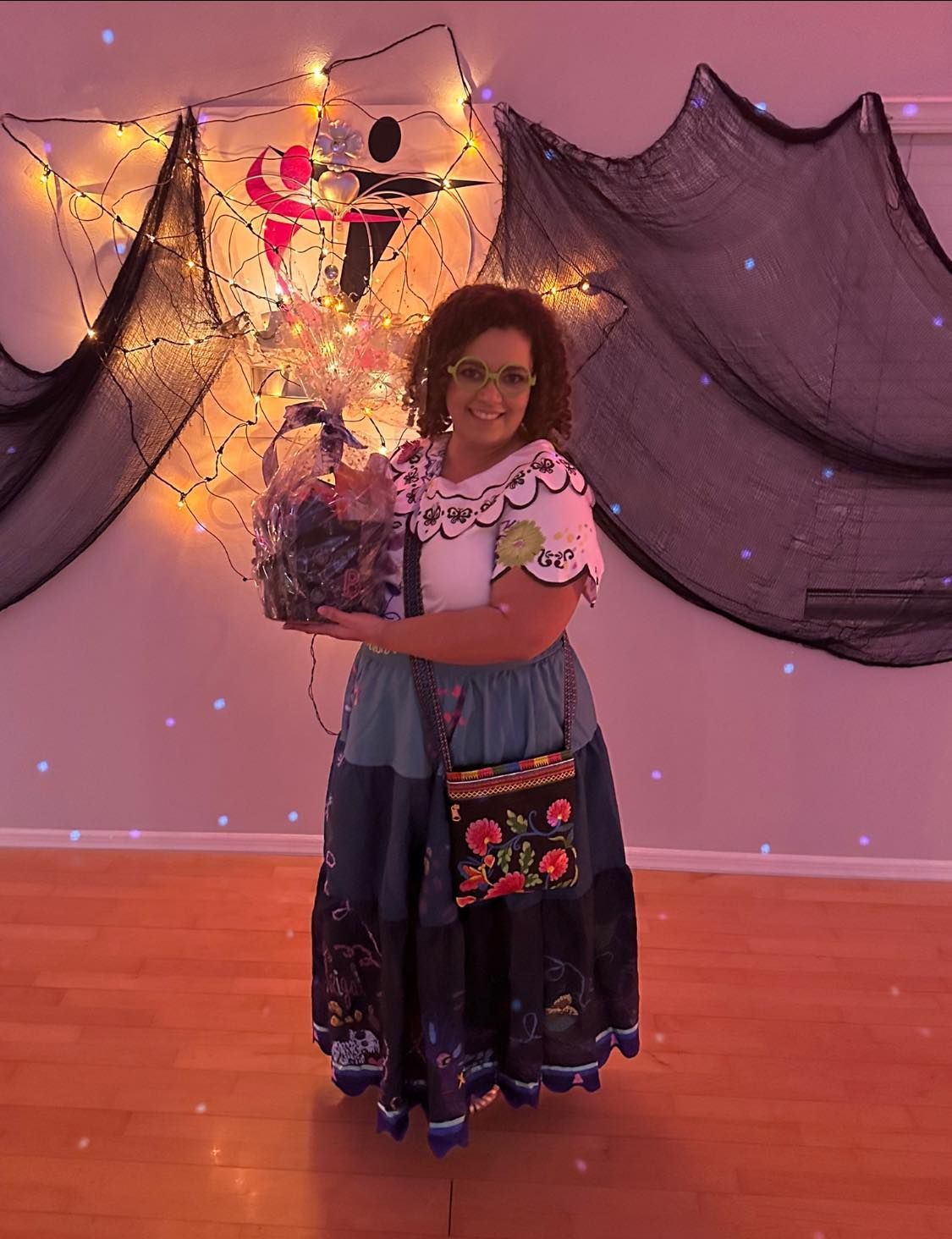 Woman in costume holding a gift basket, smiling, in front of a decorated backdrop with lights.