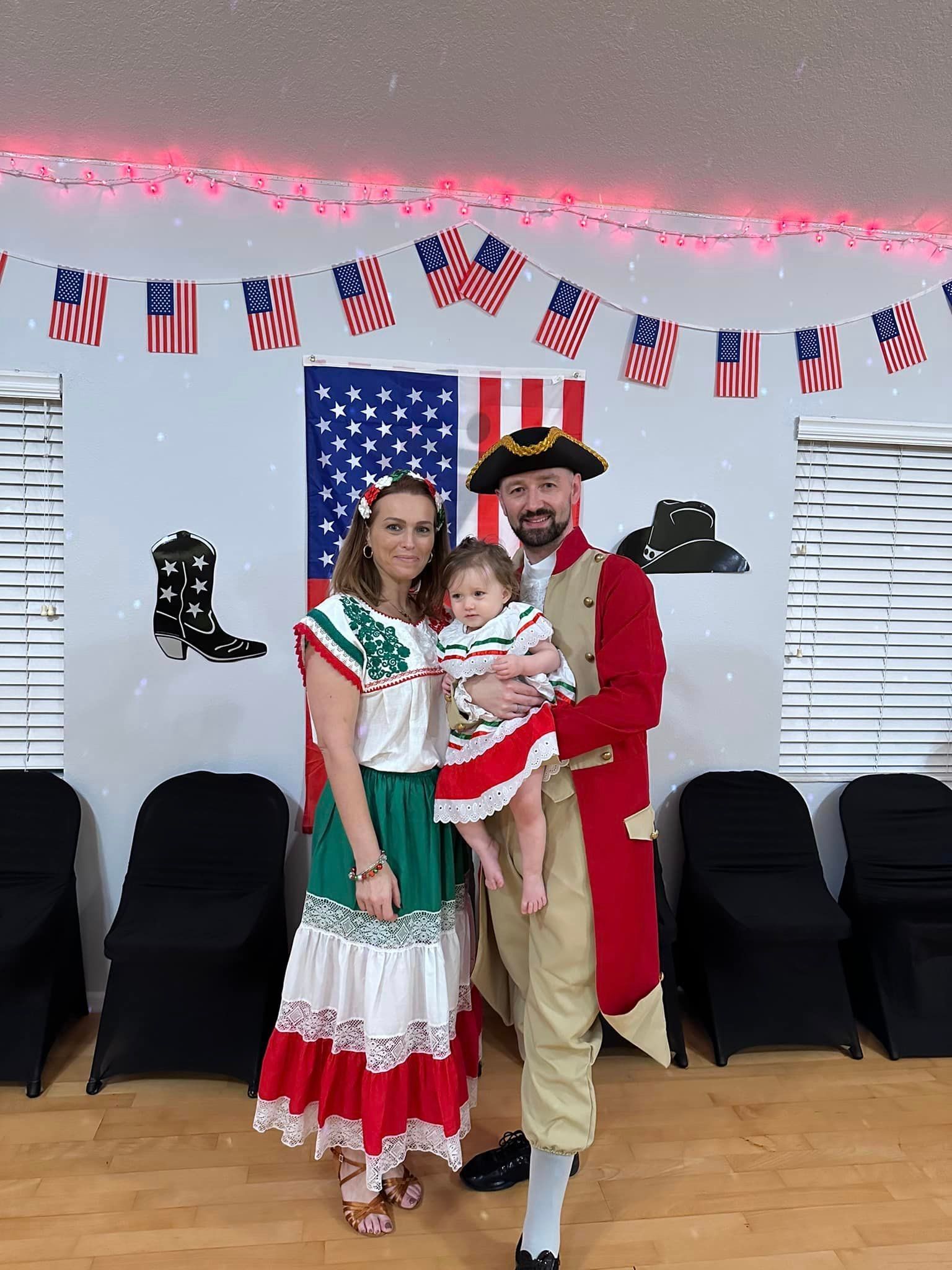 Family dressed in costumes posing in front of American flags: woman in Mexican dress, man in historical uniform, child in matching dress.