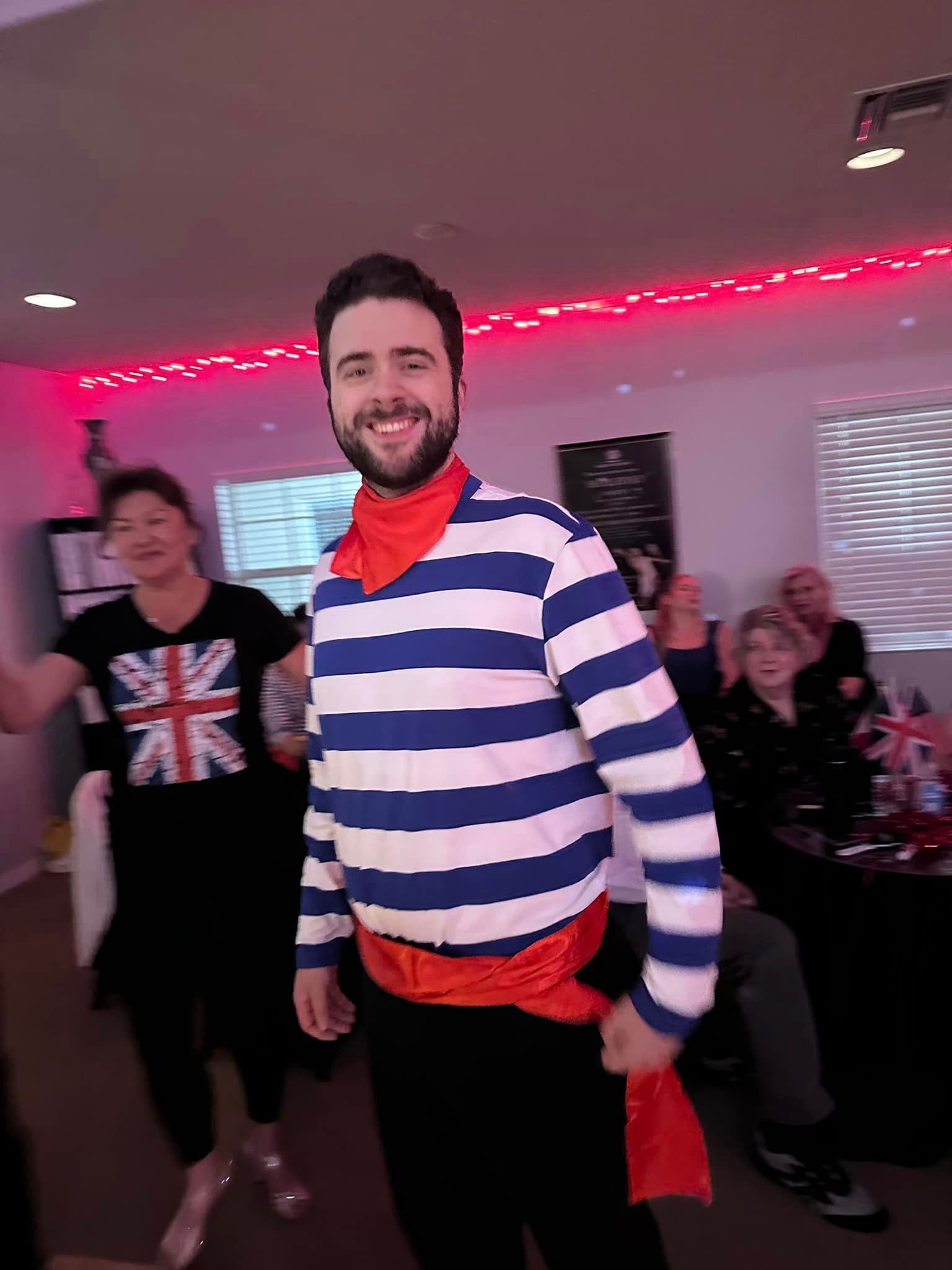 Man in striped shirt, red scarf, smiling, with others in room, celebrating.