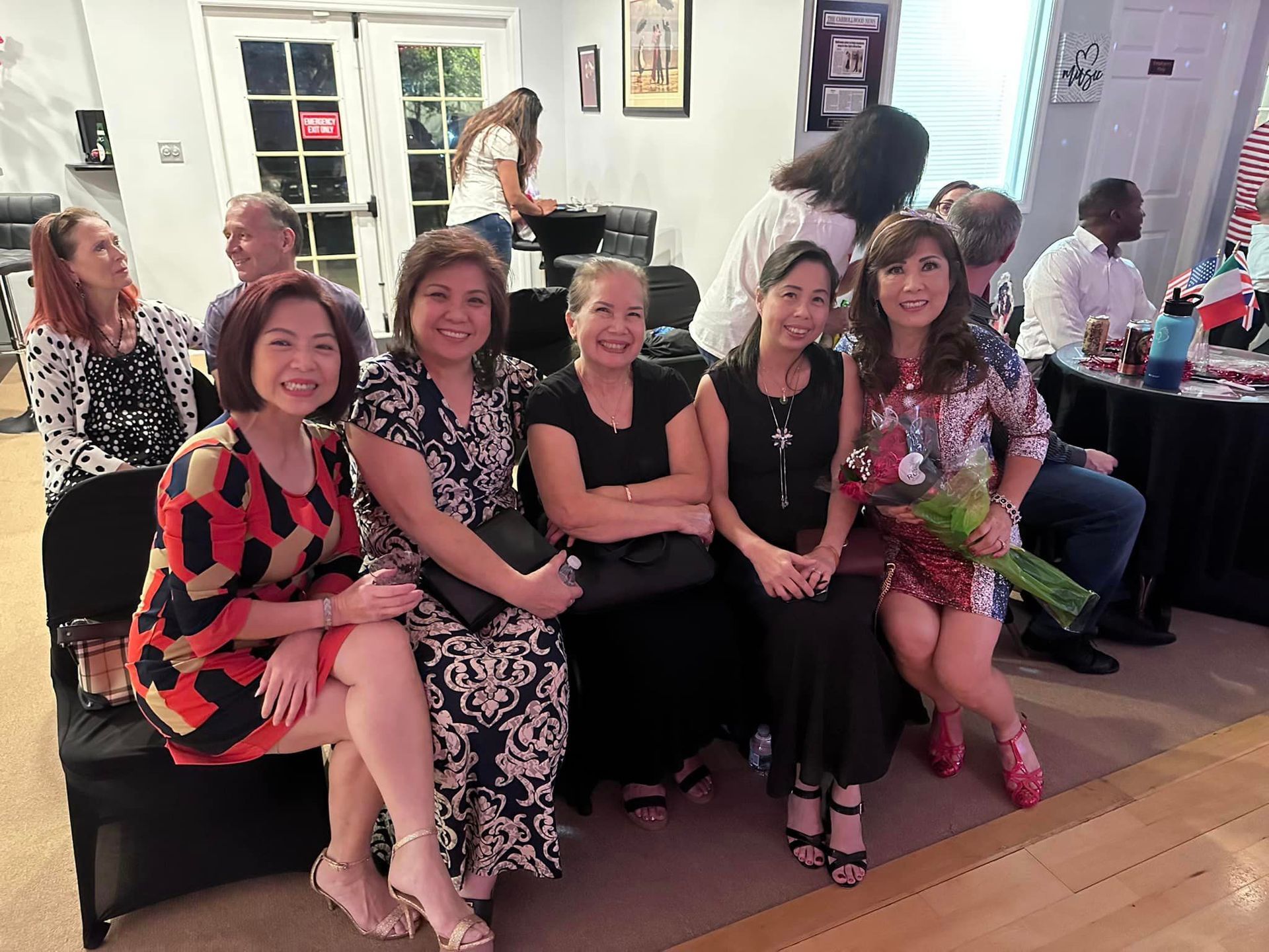 Five women smiling, posing for a photo at an event. Indoor setting with other people in the background.