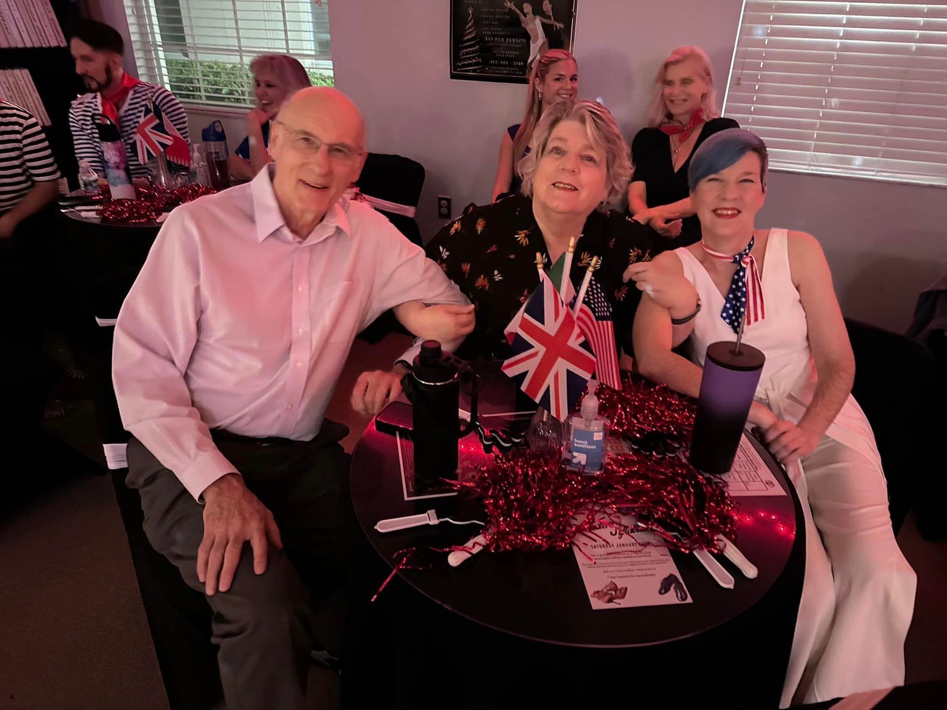 Three people at a decorated table: man, woman with crown, woman in white, all smiling, with British and American flags.