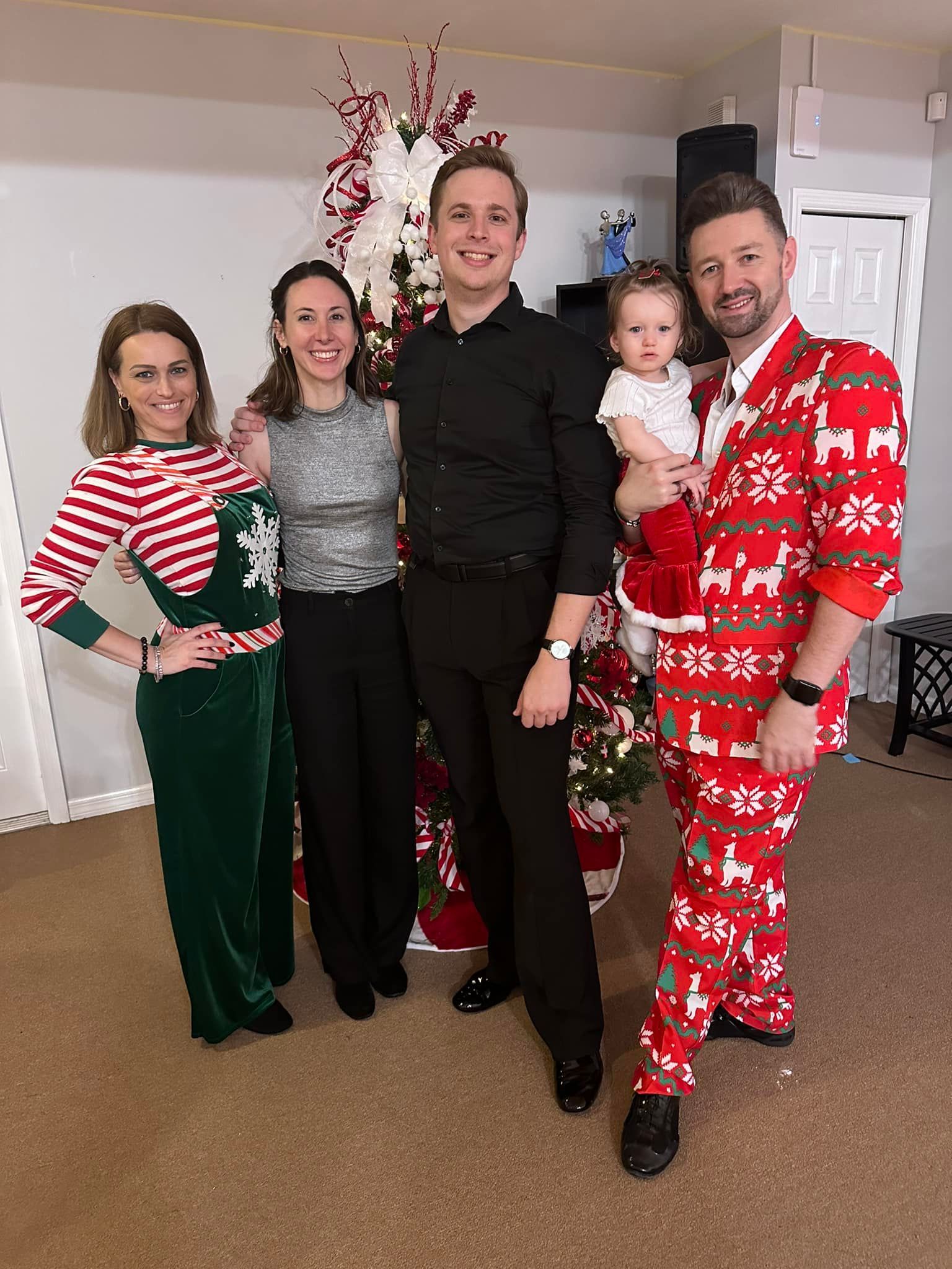 Family posing by a Christmas tree: woman in red suit, child in Santa outfit, others in festive attire.