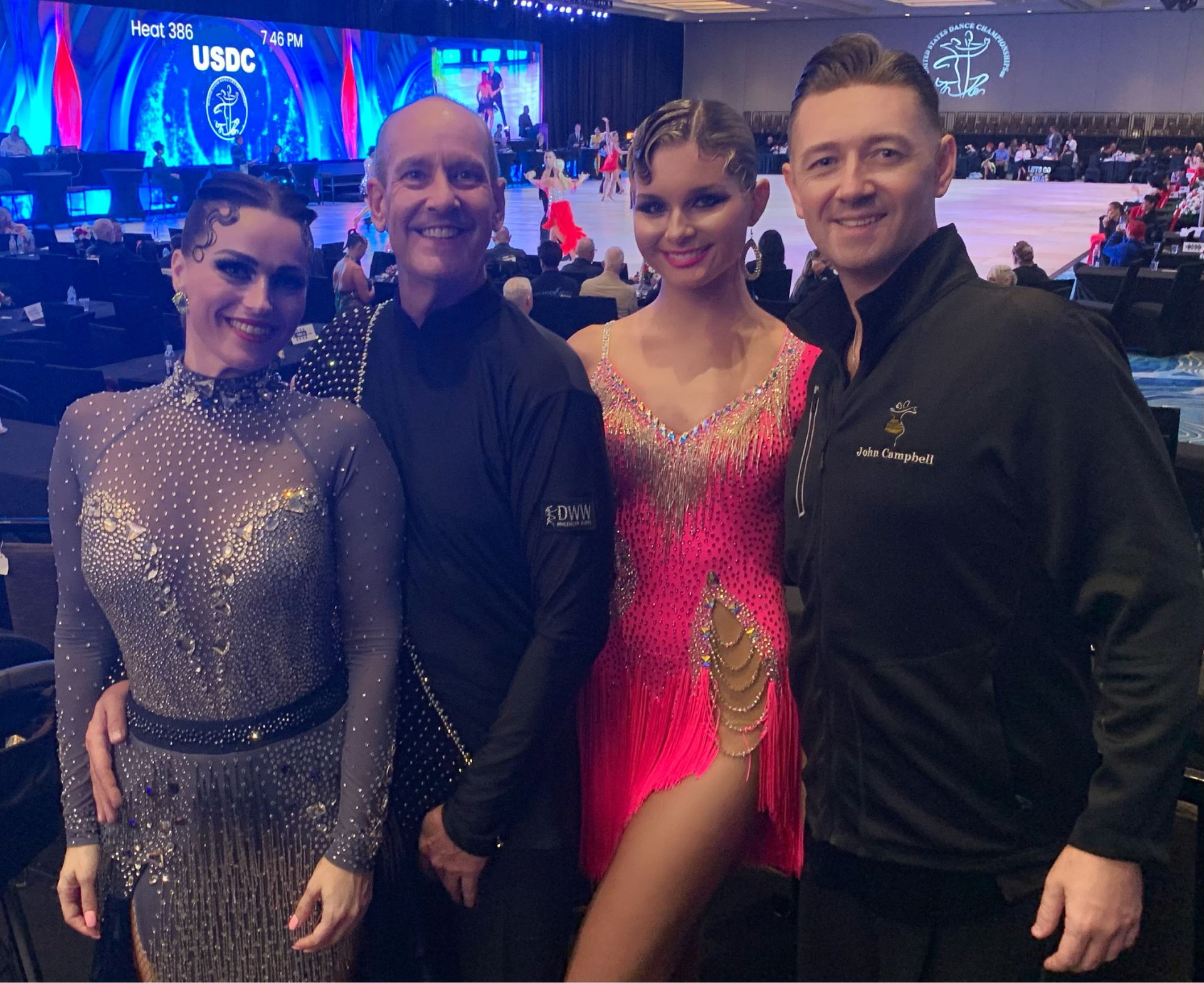 Four ballroom dancers, two couples, in a competition setting. Two women in shimmering gowns, two men in black.