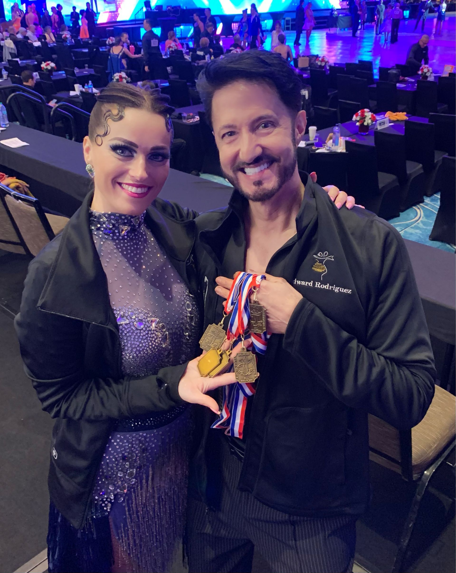 A smiling couple, a woman and a man, proudly display medals at a dance competition.