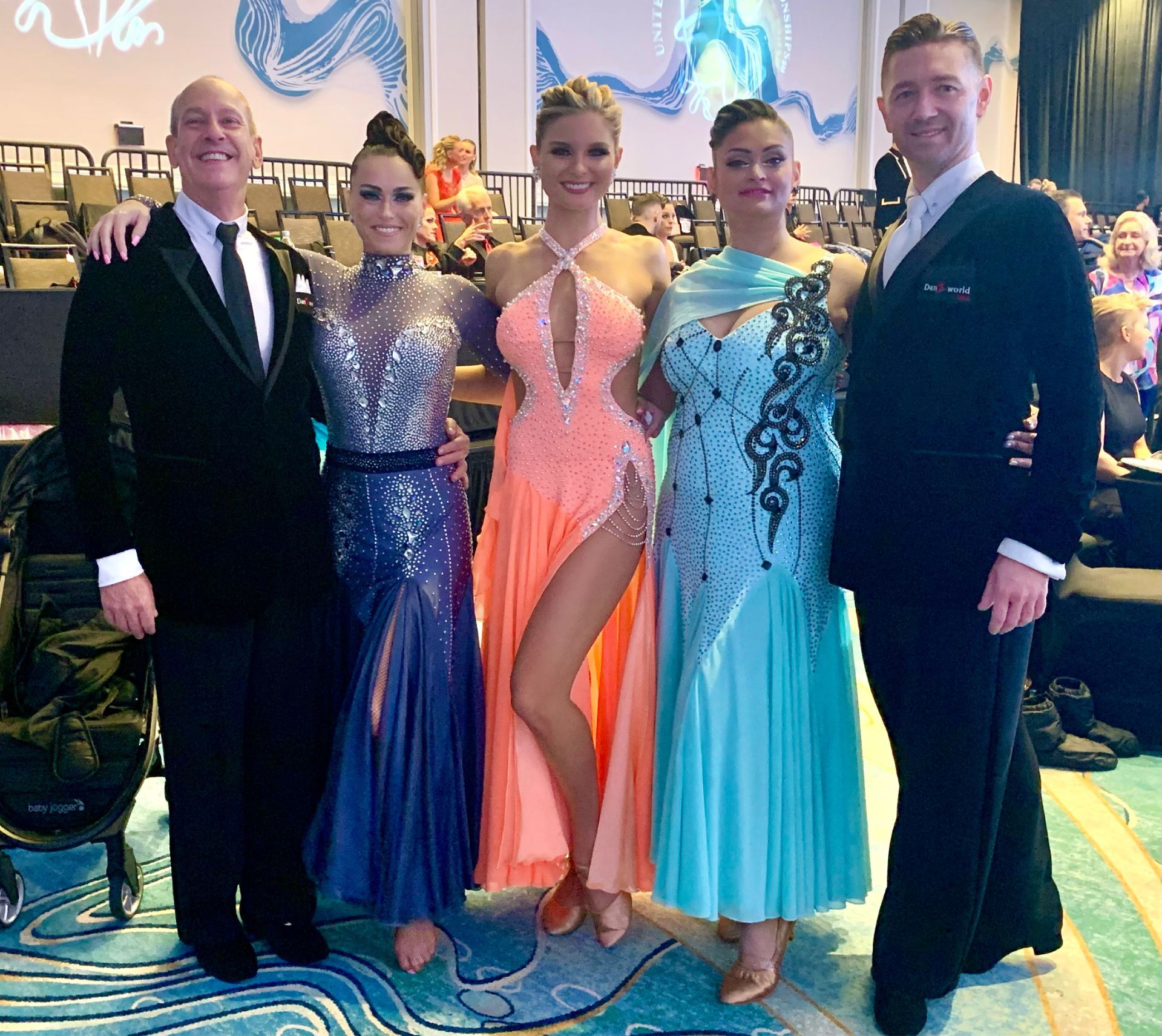 Five people in formal attire pose. Two men wear black suits, three women wear sparkly gowns, indoors.
