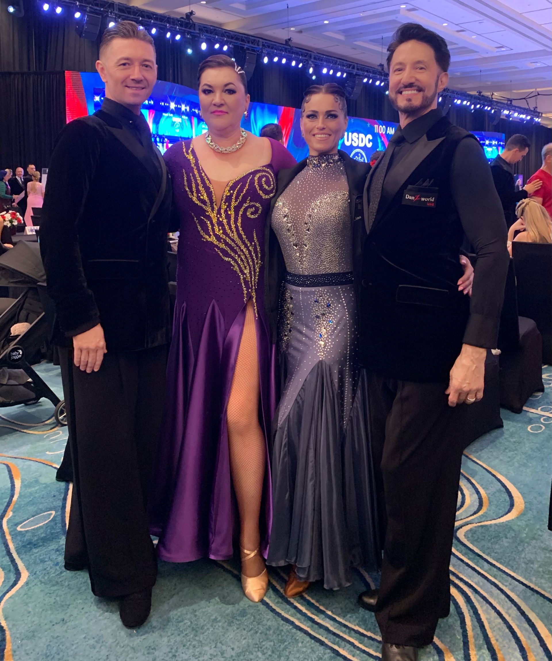 Four people in formal wear pose at an event. Two women wear gowns. Two men wear suits. Blue and red stage backdrop.