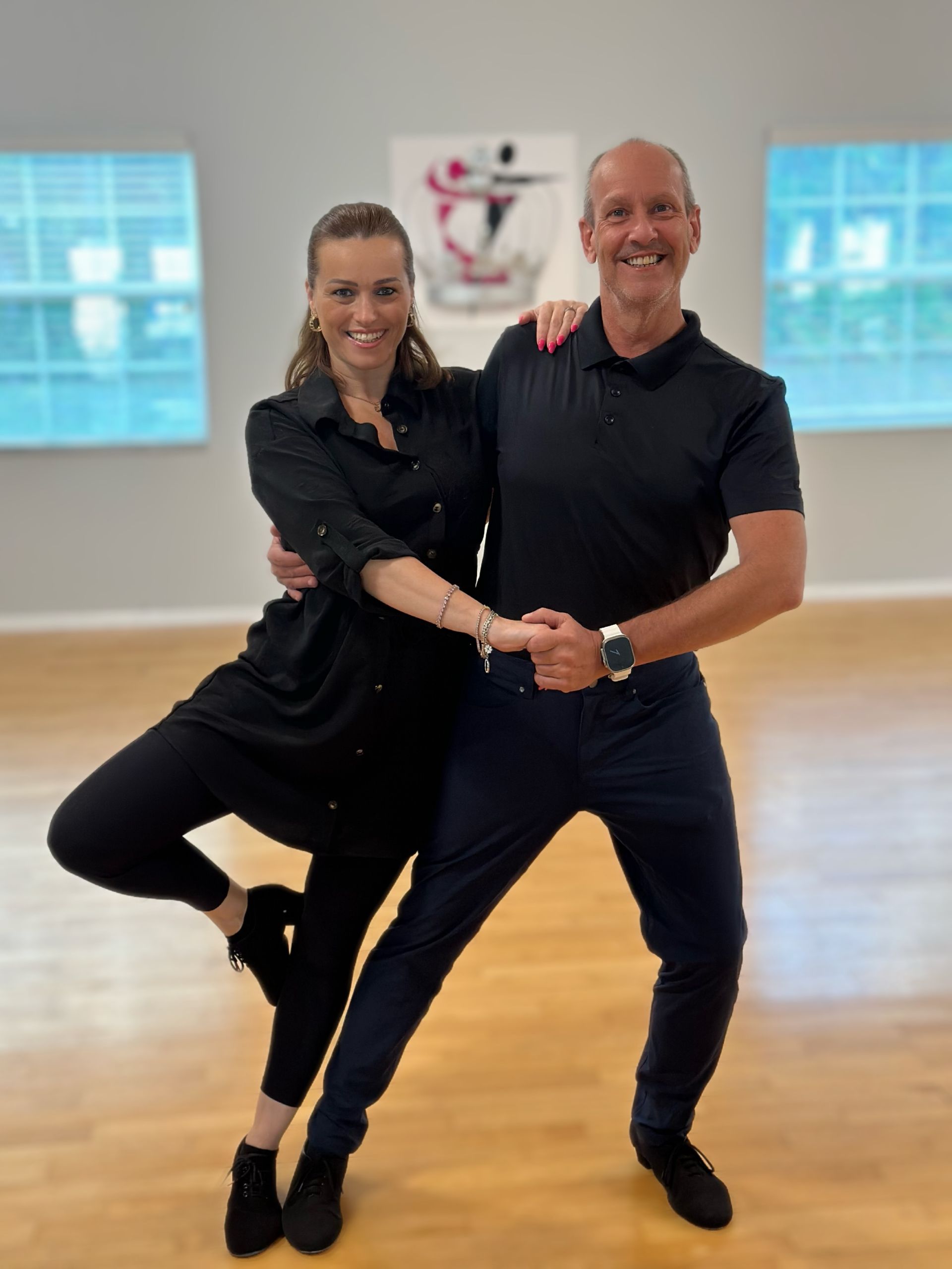 Woman and man dancing in a studio, she is wearing black and he is wearing a black shirt and dark pants.