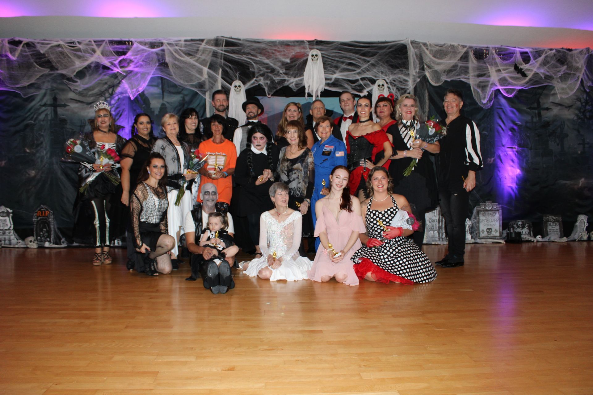 Group of people in Halloween costumes pose on a dance floor, decorated with cobwebs, ghosts, and a spooky backdrop.
