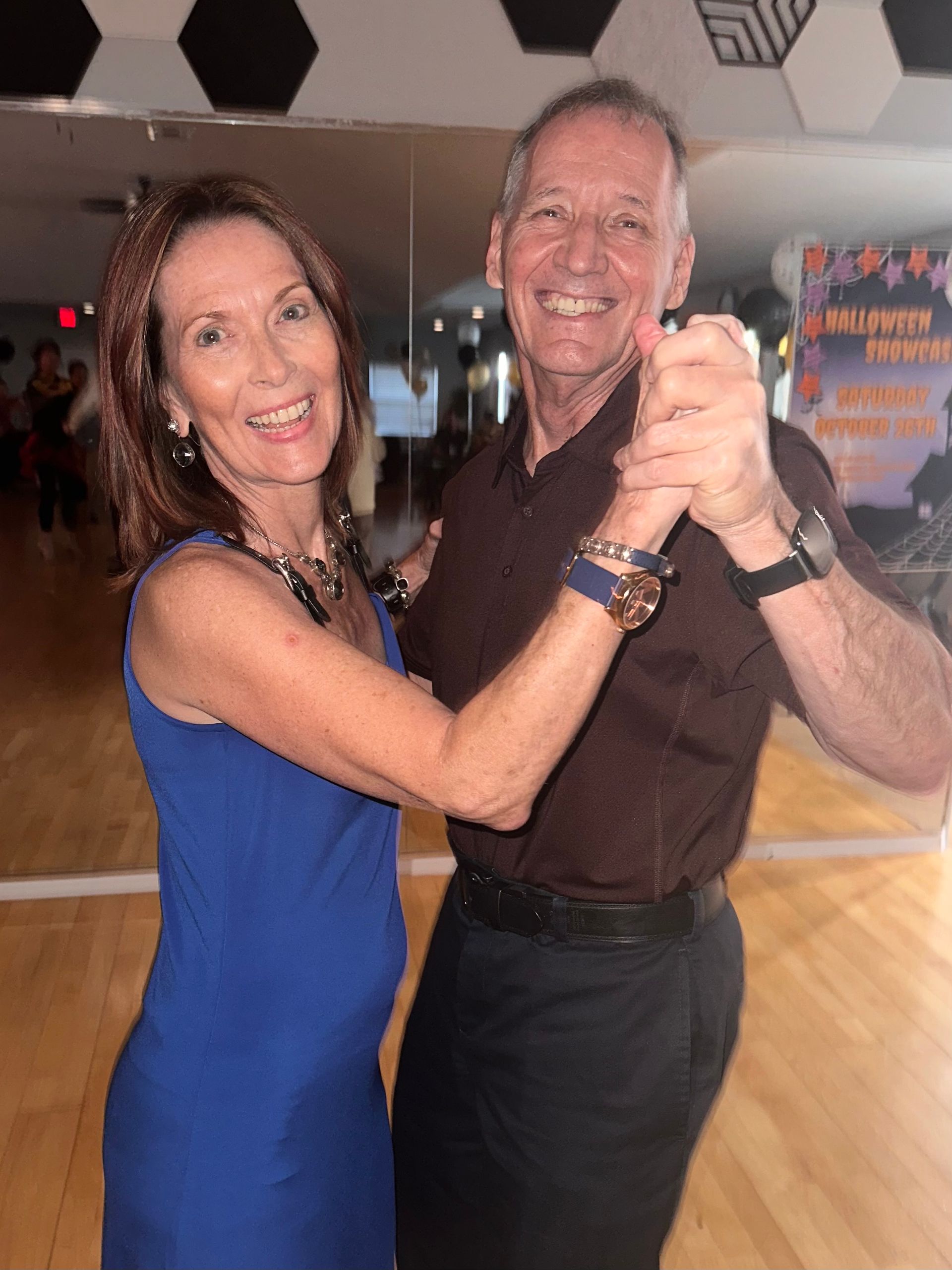 Smiling couple dancing in a studio; woman in blue dress, man in brown shirt.
