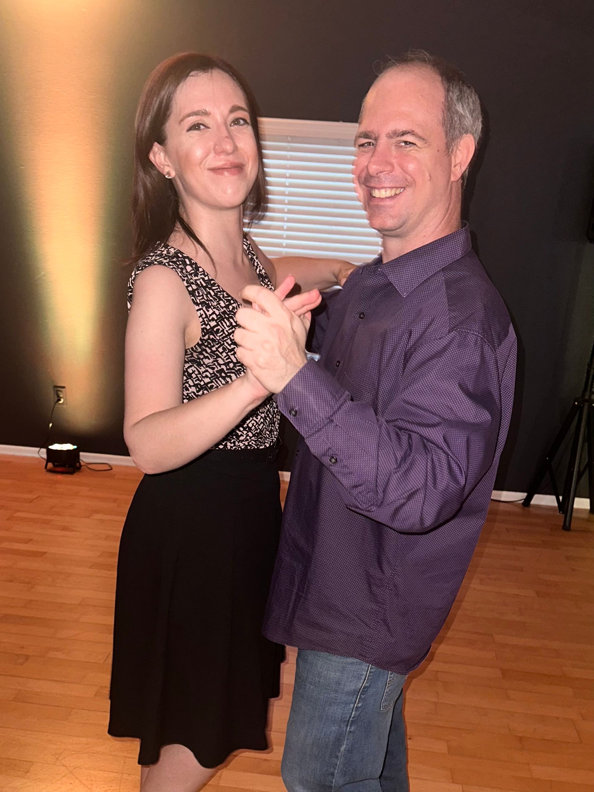Woman and man smiling while dancing in a studio; the man wears a purple shirt and jeans.
