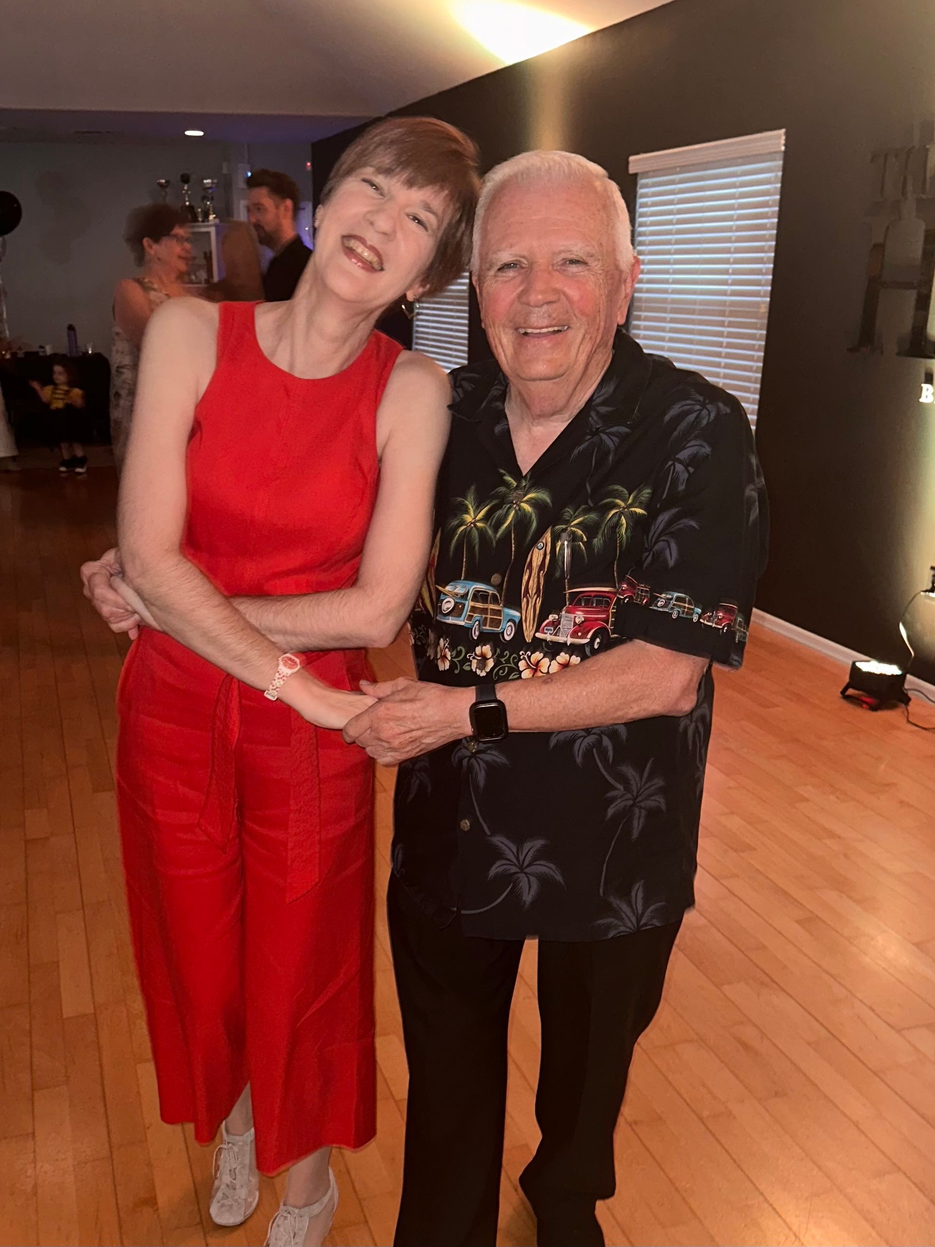 Woman in red jumpsuit and man in Hawaiian shirt smiling and holding hands.