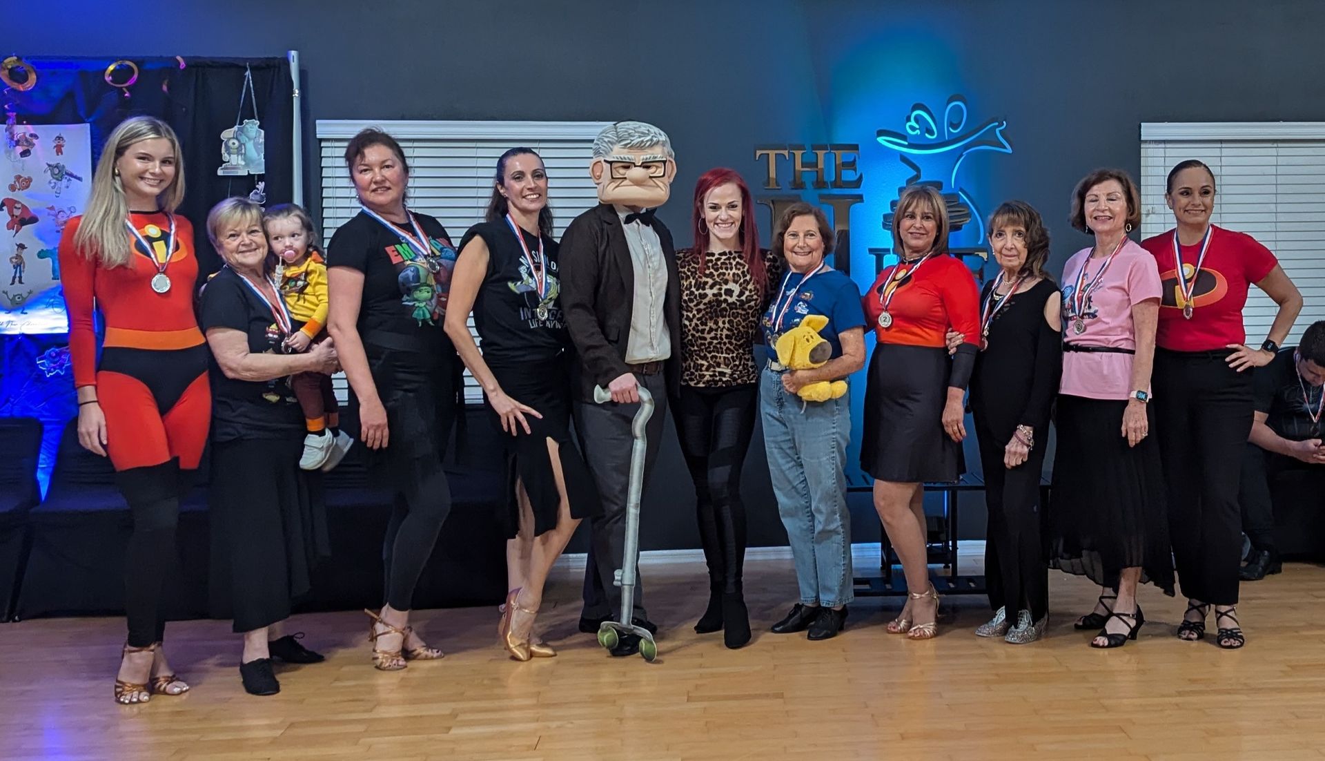 Group posing at a dance studio; diverse people in costumes, some with medals, smiling.