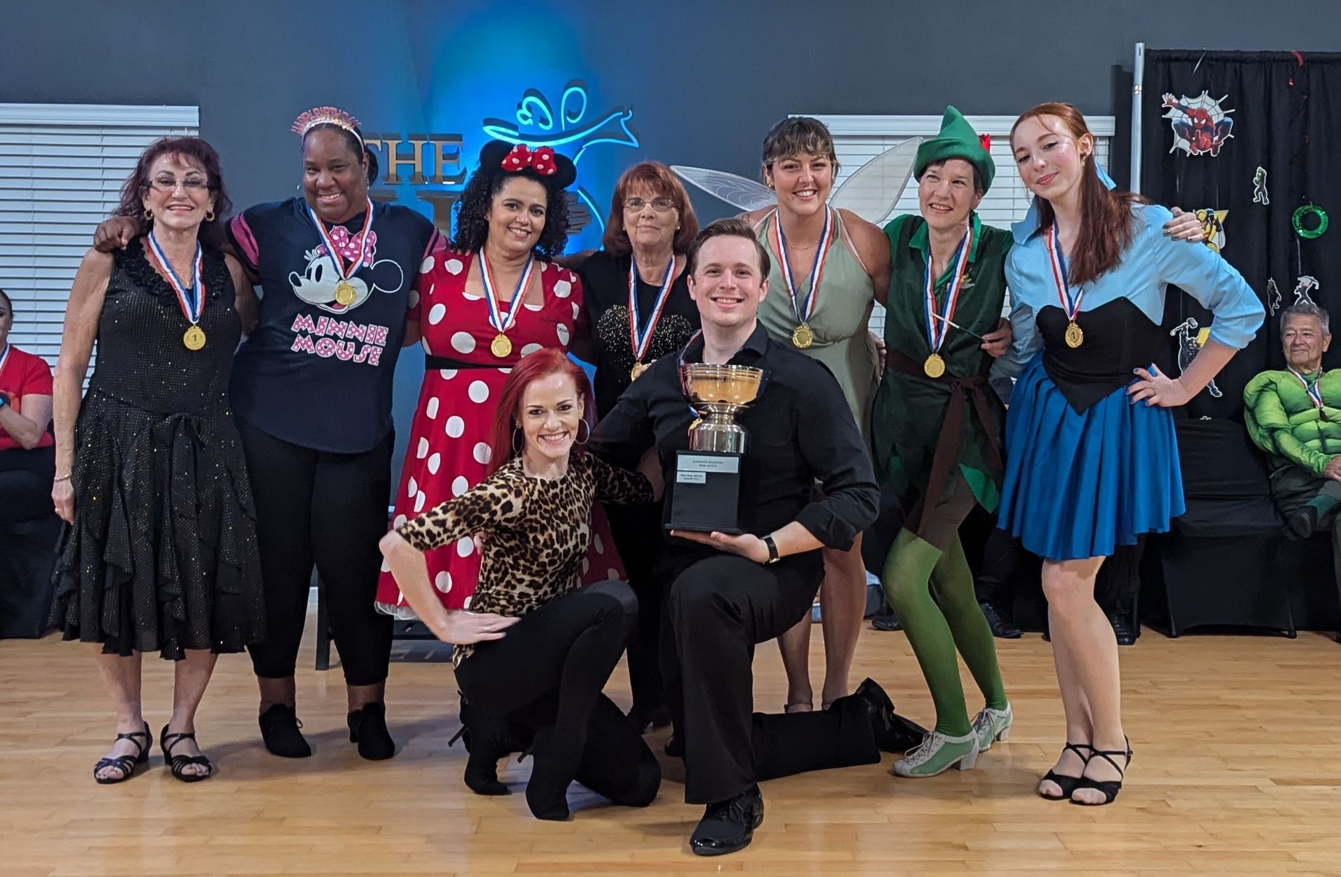 Group of dancers in costume with medals posing for a photo. Man holds a trophy in a dance studio.