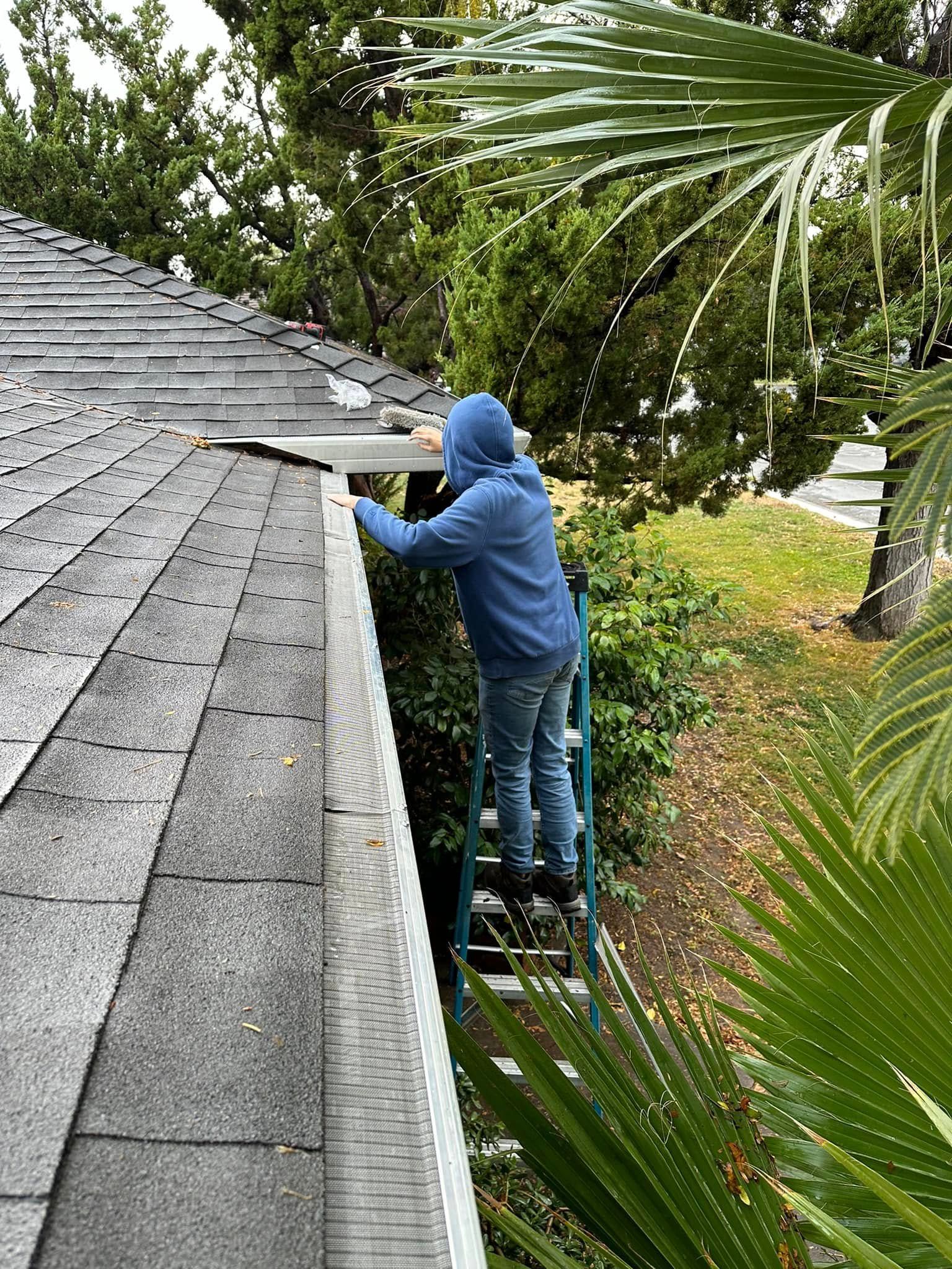 Person in a blue hoodie working on a shingled roof beside trees and a ladder