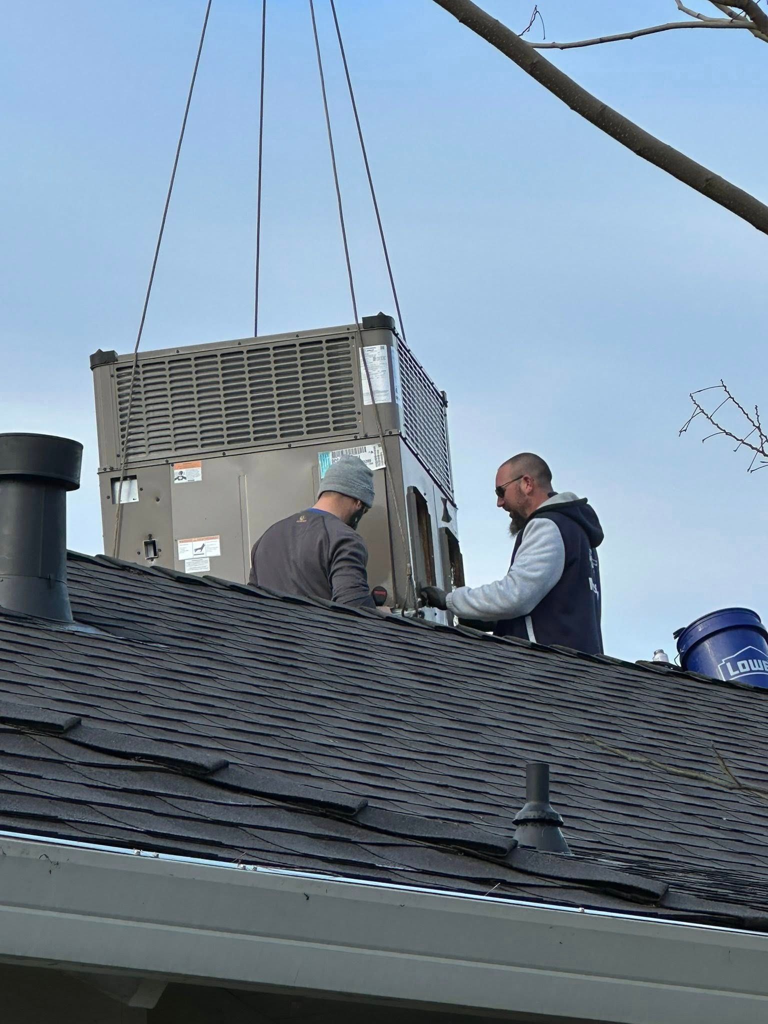 Two workers on a shingled roof lifting an HVAC unit with a crane