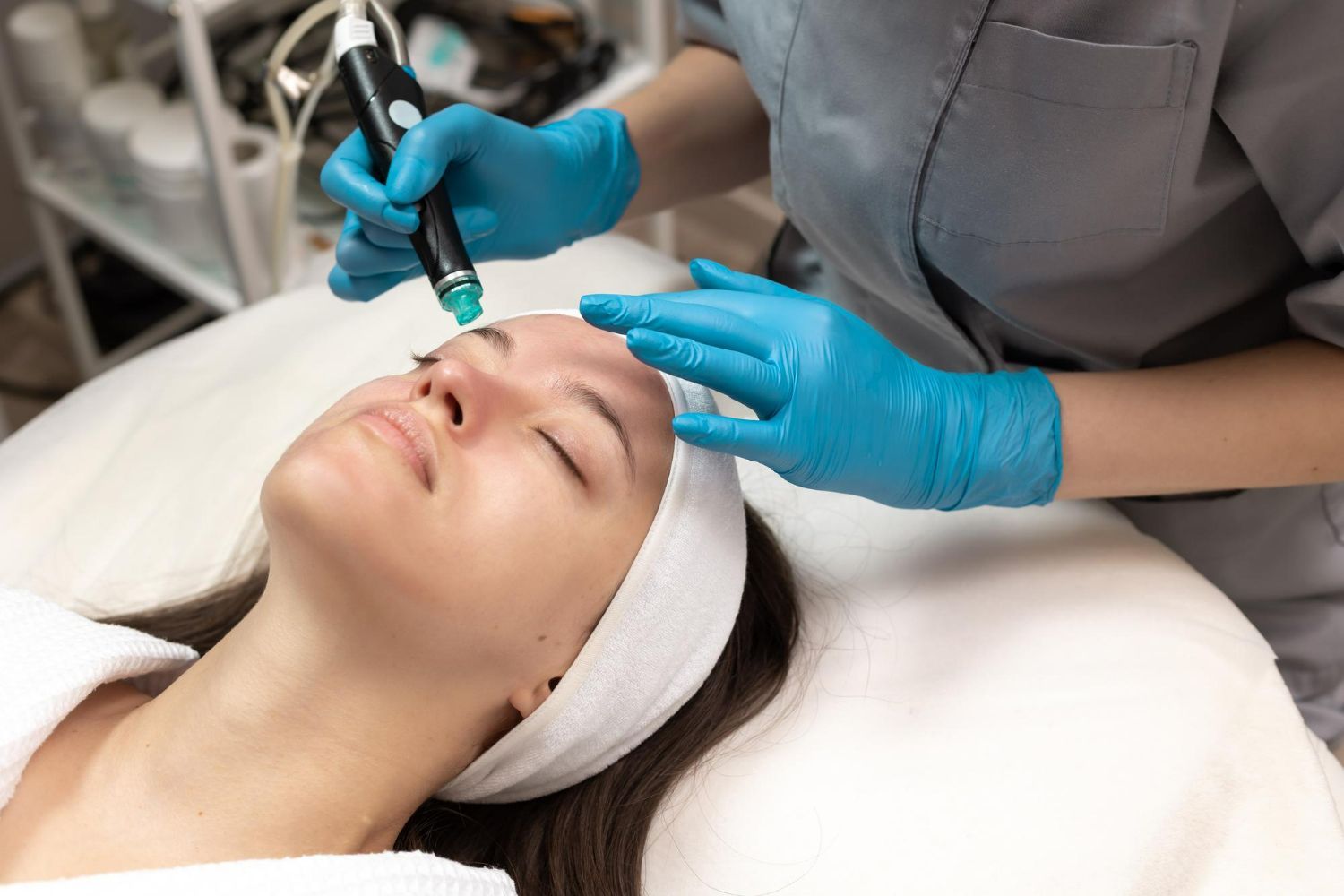 Person receiving facial treatment with tool, in a spa setting. The practitioner wears blue gloves.