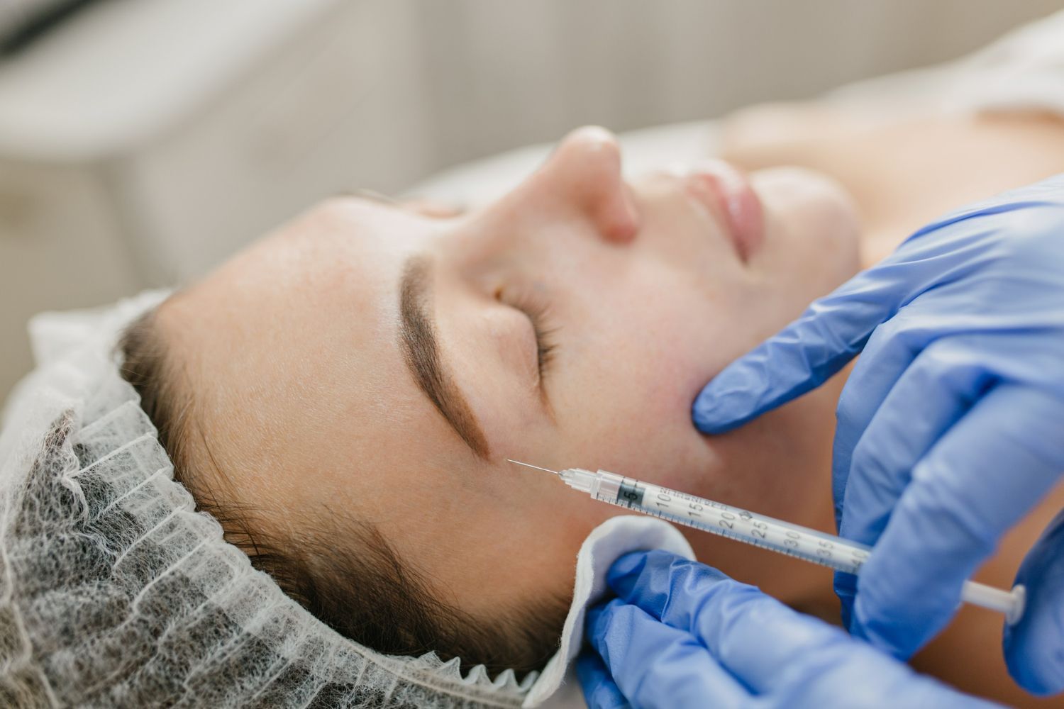 Person receiving facial injection in a medical setting. Blue gloves, needle, white cap.