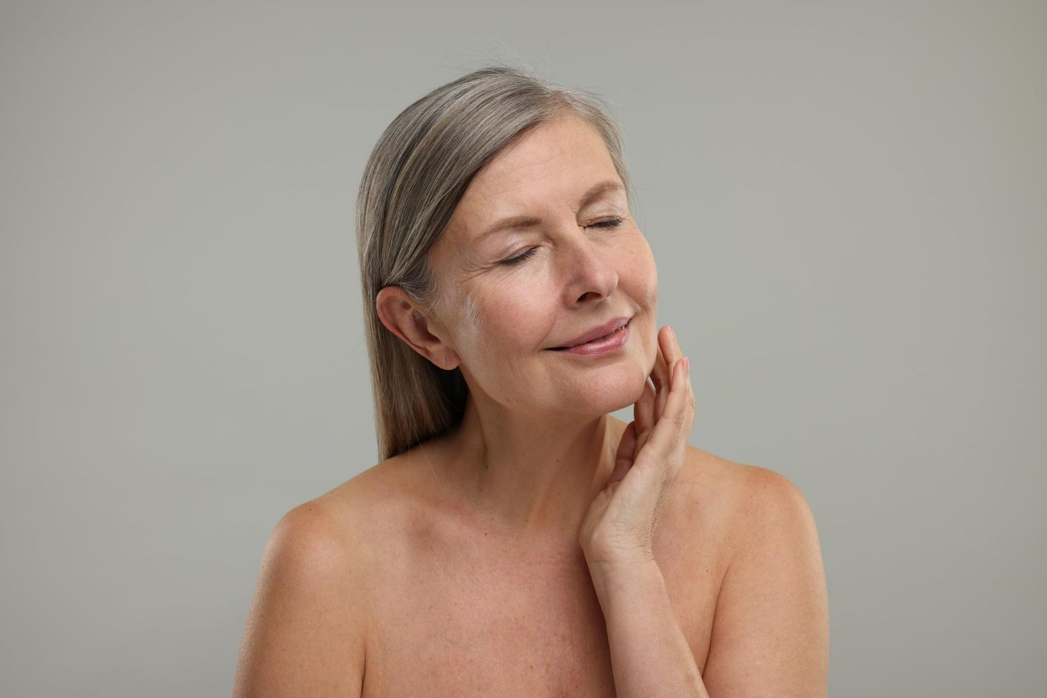 Woman with gray hair and closed eyes touches her neck, smiling. Neutral background.