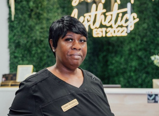 A woman in a black scrub top is standing in front of a sign that says aesthetics est 2022.