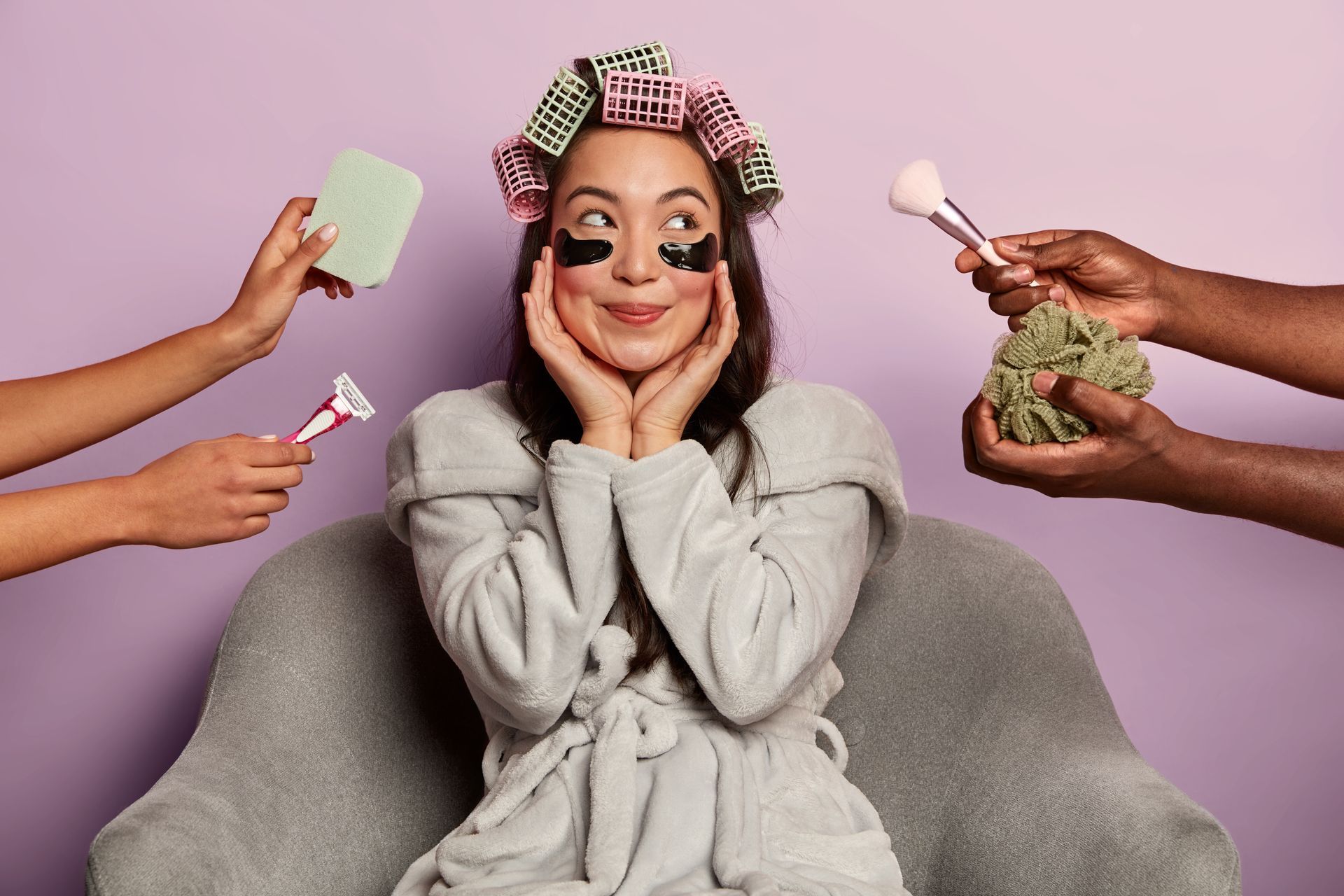 A woman with curlers in her hair is sitting in a chair surrounded by people holding cosmetics.