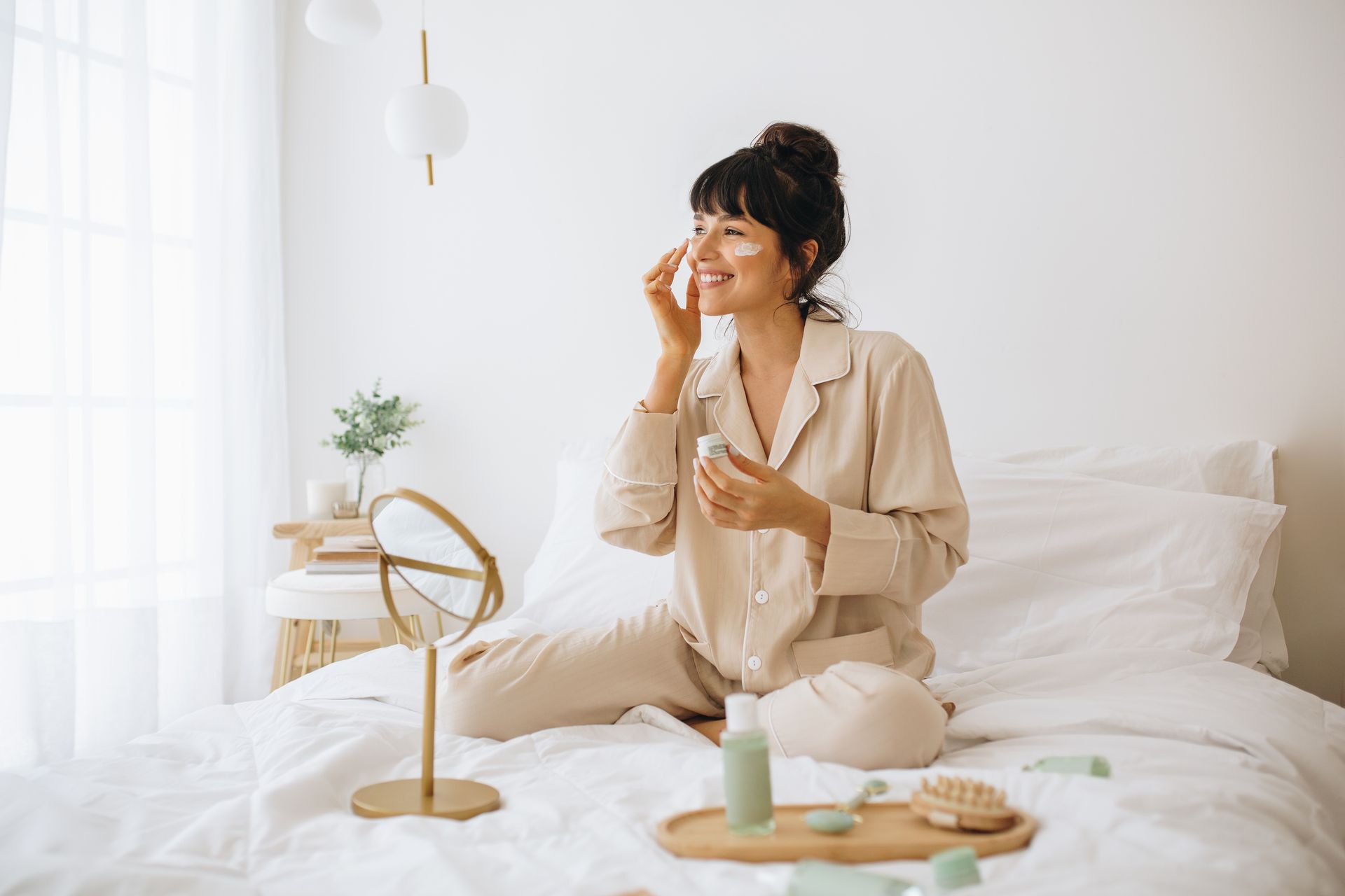 A woman is sitting on a bed applying lotion to her face.