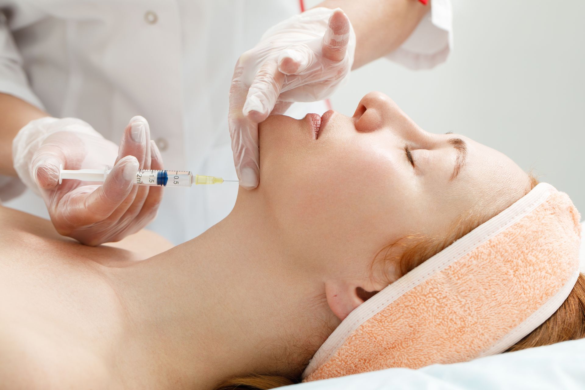 Woman receiving a cosmetic injection in her jawline; hands with gloves, syringe, setting is a clinic.