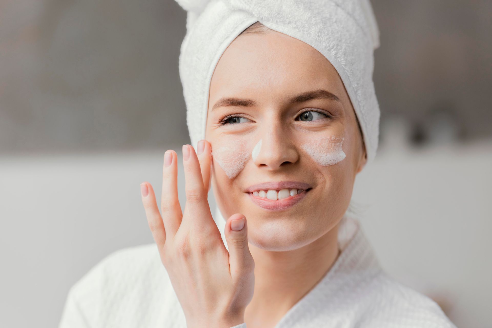 A woman with a towel wrapped around her head is applying lotion to her face.