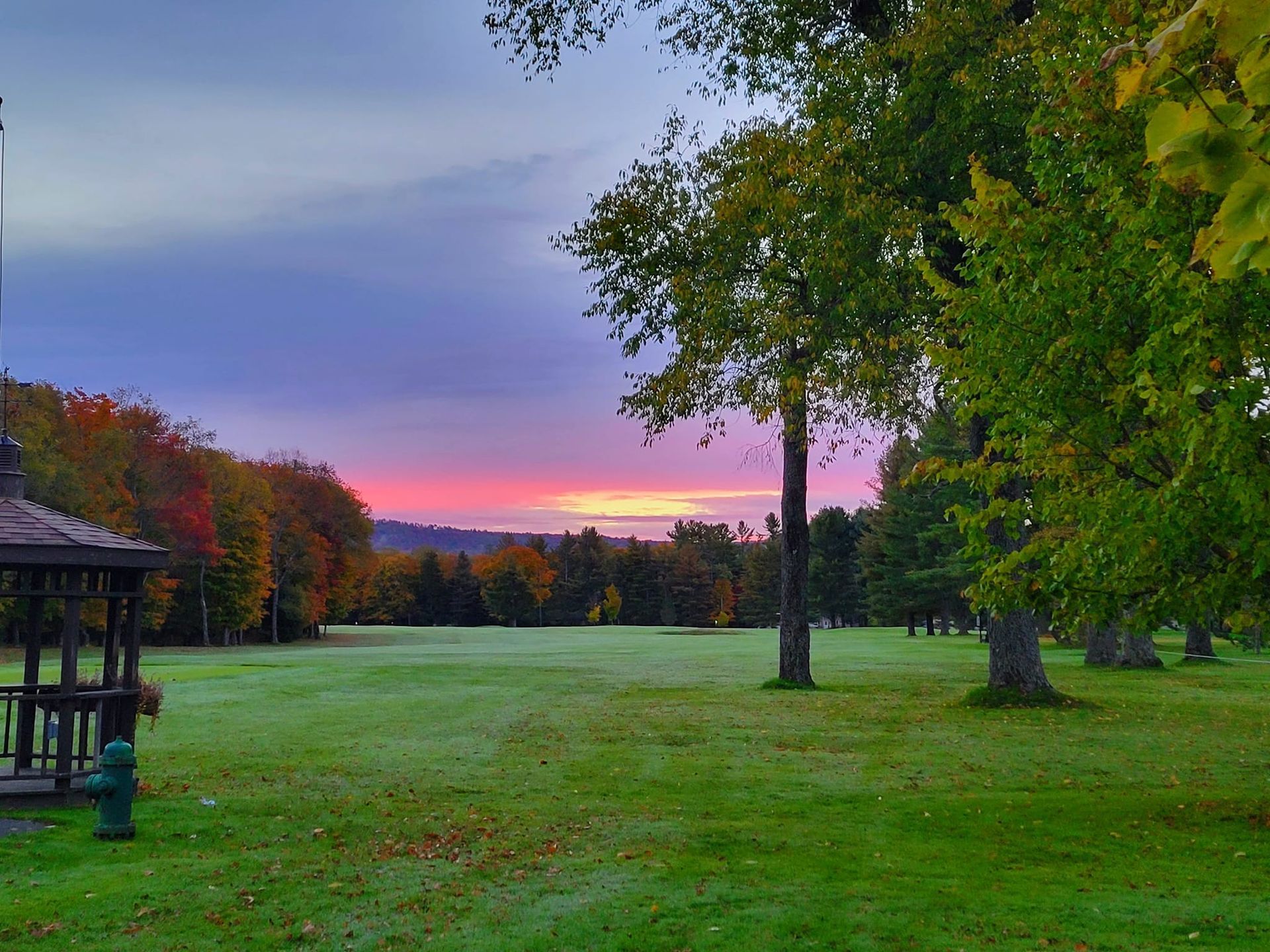 A sunrise glows pink and orange above a green field with trees and a gazebo, creating a peaceful, autumnal park landscape.