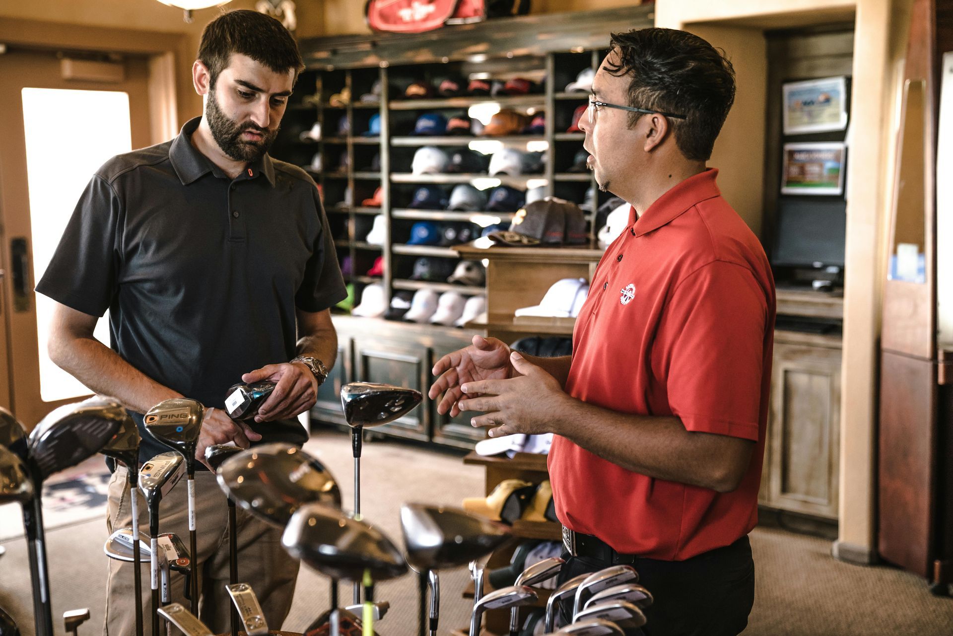Two men stand in a golf pro shop, one in a red shirt talking while the other in a grey shirt examines a golf club.