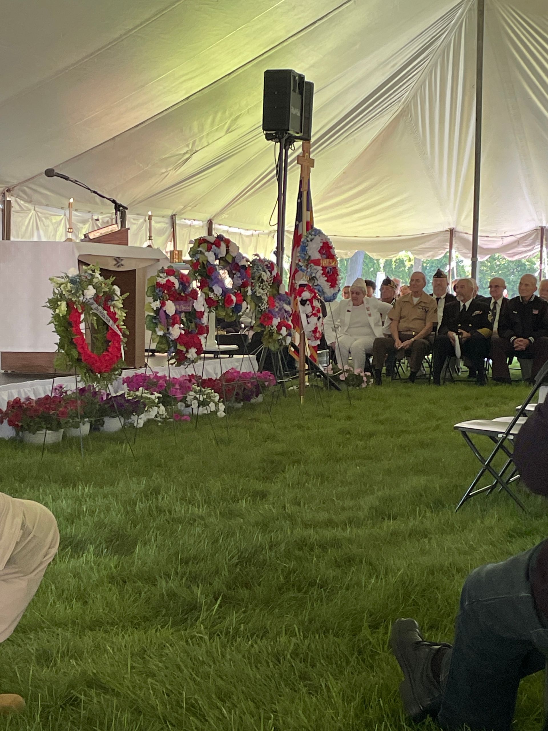 Memorial service under a white tent with flower wreaths, seated attendees, and a speaker on stage