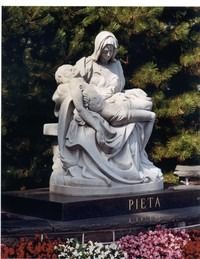 White Pieta statue on a dark pedestal, with flowers in front and trees behind.